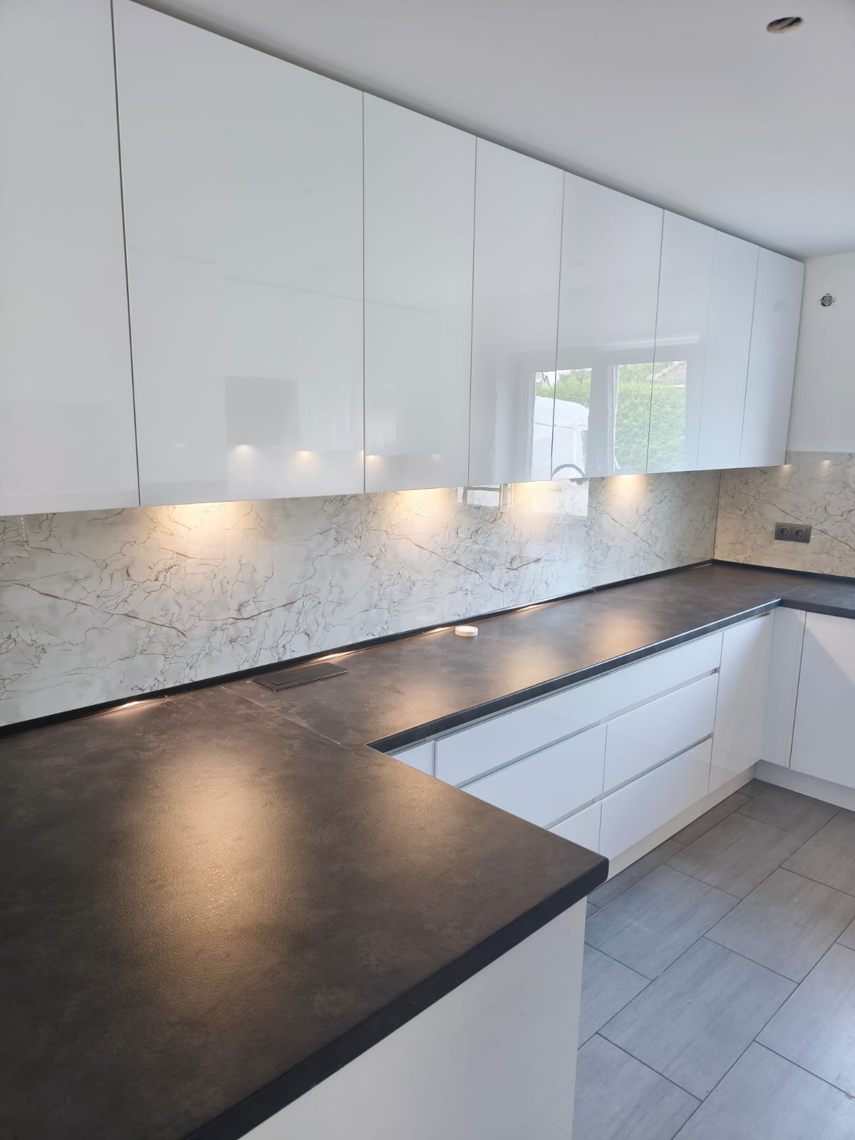 Biała, nowoczesna kuchnia z połyskiem, ciemny blat i marmurowy panel nad blatem, oświetlenie LED podszafkowe, szare płytki podłogowe. Minimalistyczny design.