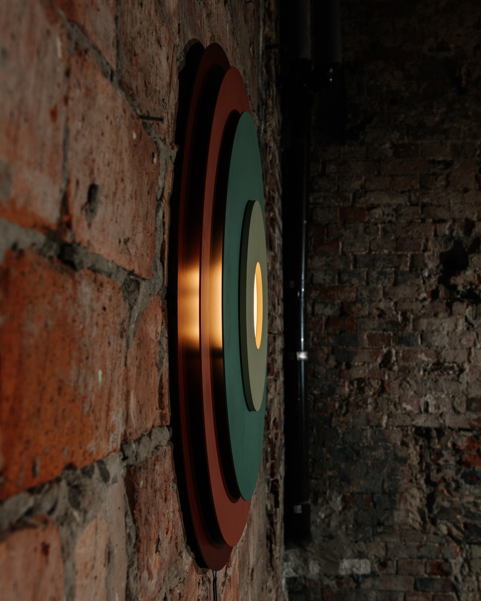 Dekoracyjna lampa ścienna z okręgami w kolorach: ceglastym, zielonym i beżowym, zamontowana na ścianie z czerwonej cegły. Widoczne przewody.