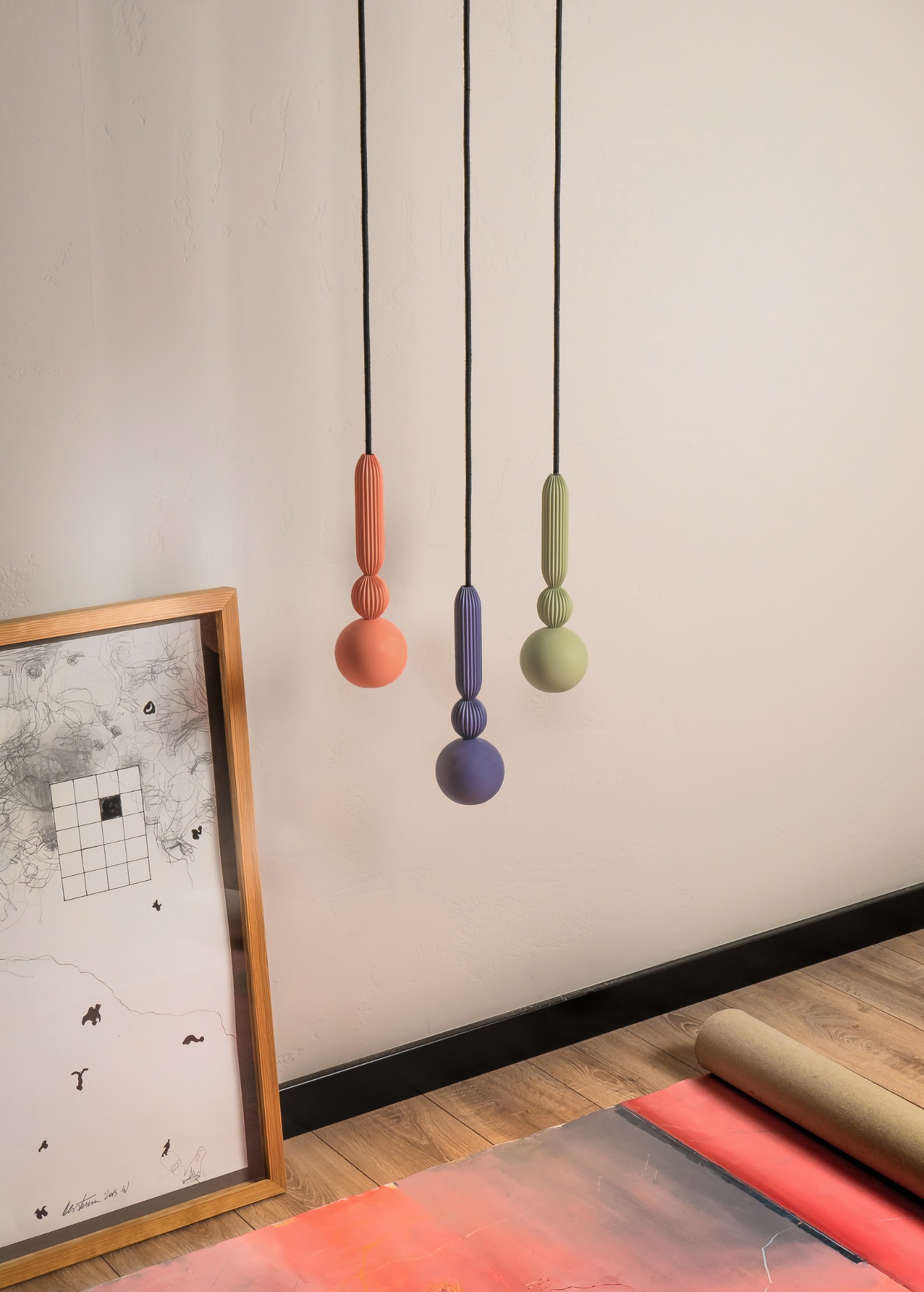 Trzy wiszące lampy o geometrycznych kształtach w pastelowych kolorach: łososiowy, fioletowy i pistacjowy. Obok obraz w drewnianej ramie i zwinięty plakat na podłodze.