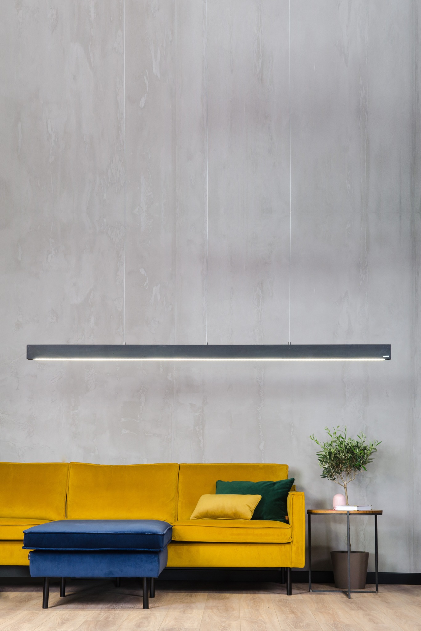Nowoczesna, długa lampa wisząca LED w szarym kolorze, oświetlająca żółtą sofę i niebieski puf na tle ściany z efektem betonu. Minimalistyczny design.