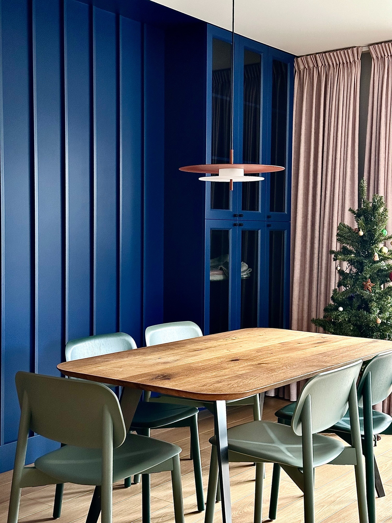 Nowoczesna jadalnia z drewnianym stołem i krzesłami w odcieniach zieleni, oświetlona designerską lampą wiszącą. W tle niebieska ściana i świąteczna choinka.