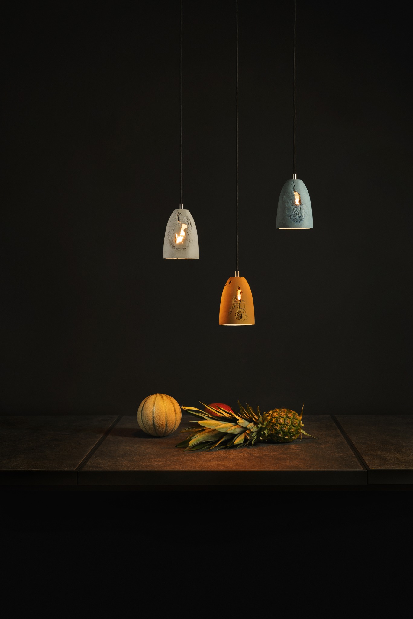 Trzy lampy wiszące o różnych kolorach (biały, pomarańczowy, niebieski) z dekoracyjnymi wzorami, umieszczone nad blatem z owocami (melon i ananas).
