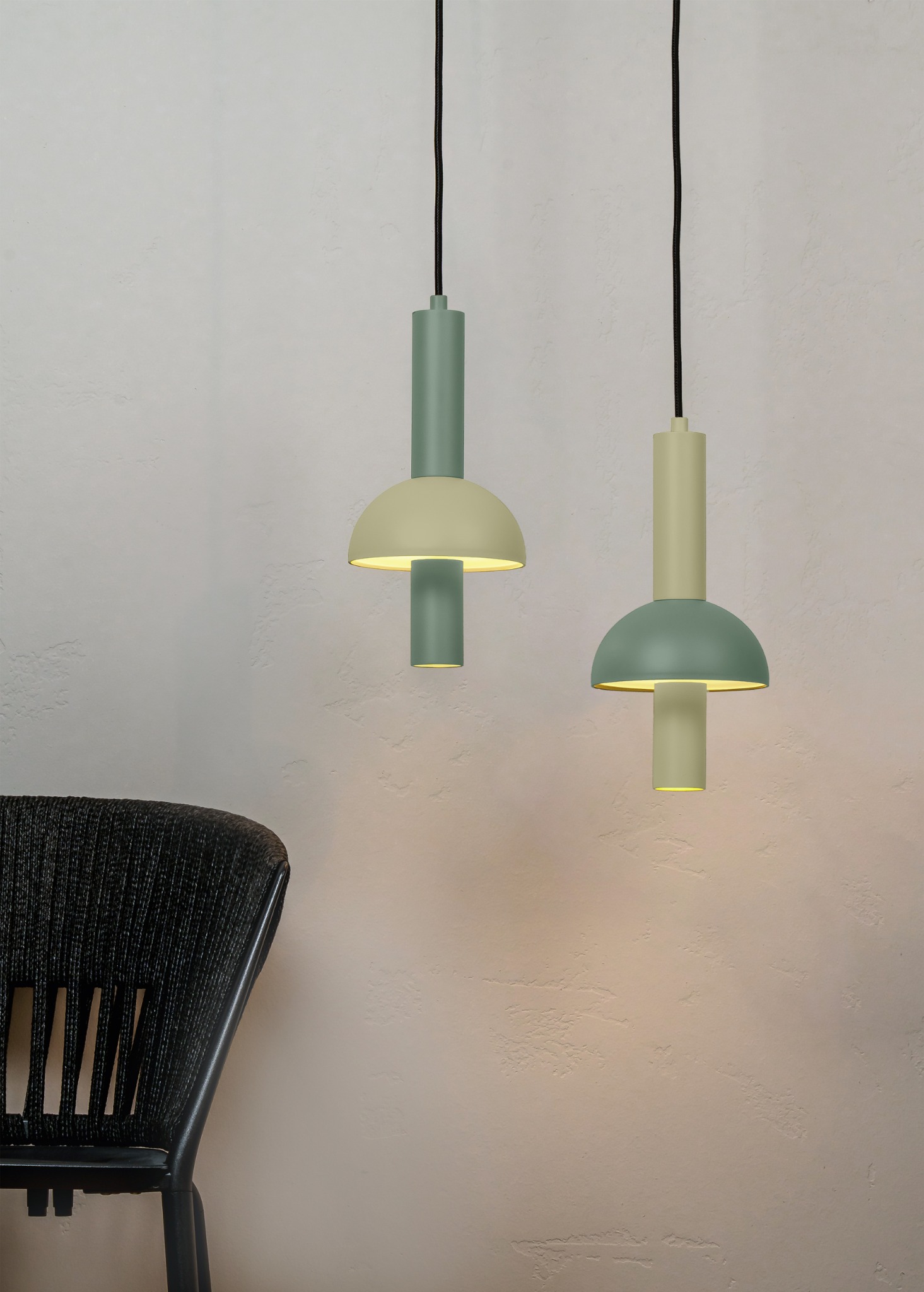 Dwa nowoczesne, pastelowe żyrandole wiszące na tle teksturowanej ściany, z fragmentem czarnego krzesła w lewym dolnym rogu kadru. Oświetlenie wnętrza.