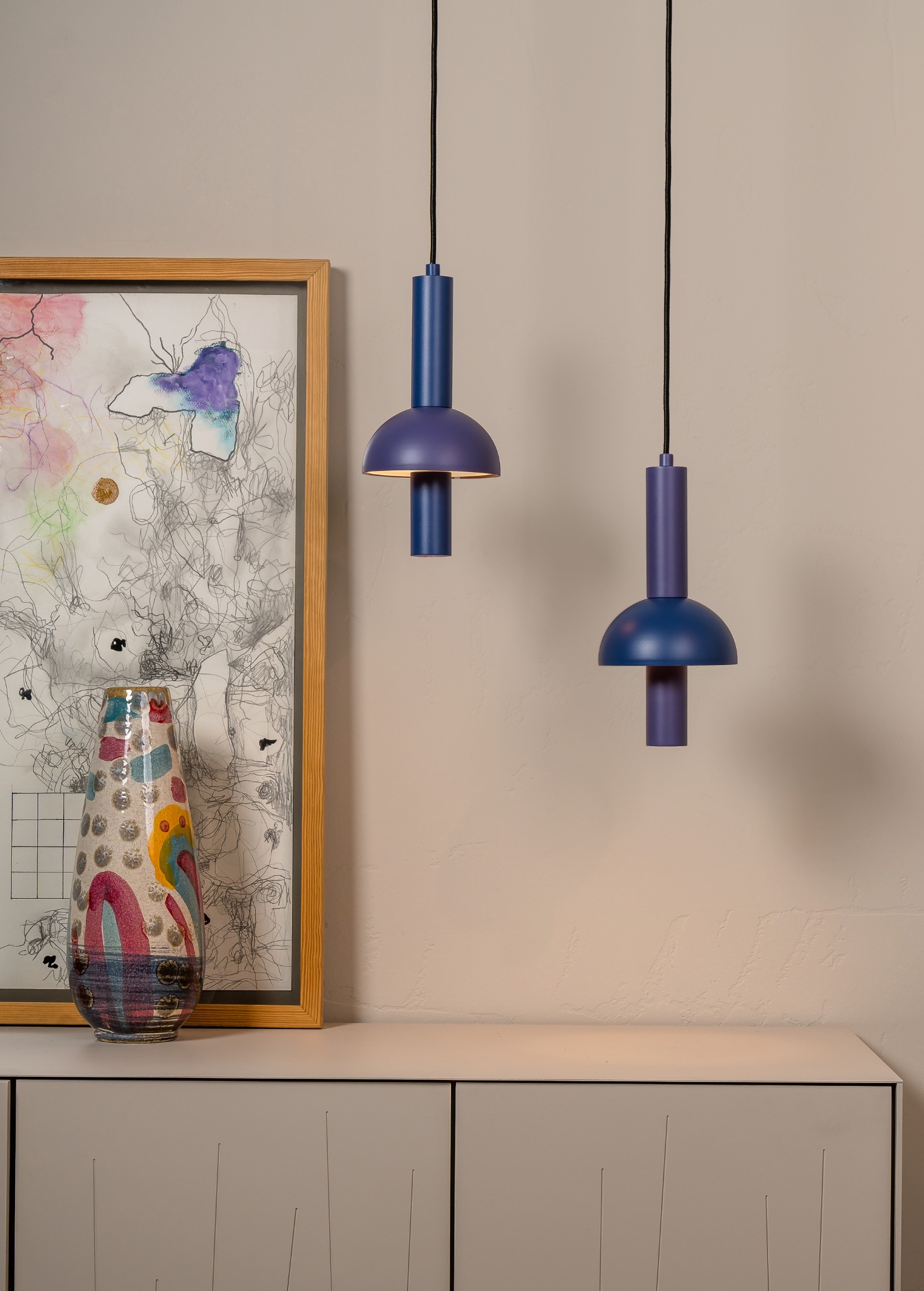 Dwie wiszące lampy w odcieniach fioletu nad komodą z dekoracjami: abstrakcyjny obraz w ramie i wzorzysty wazon. Nowoczesny design oświetlenia wnętrza.