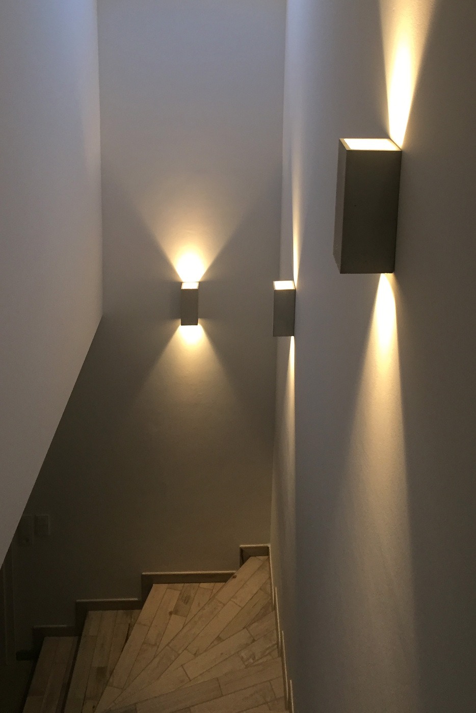 Nowoczesne oświetlenie schodów z trzema kwadratowymi kinkietami emitującymi światło w górę i w dół. Drewniane schody w dolnej części kadru.