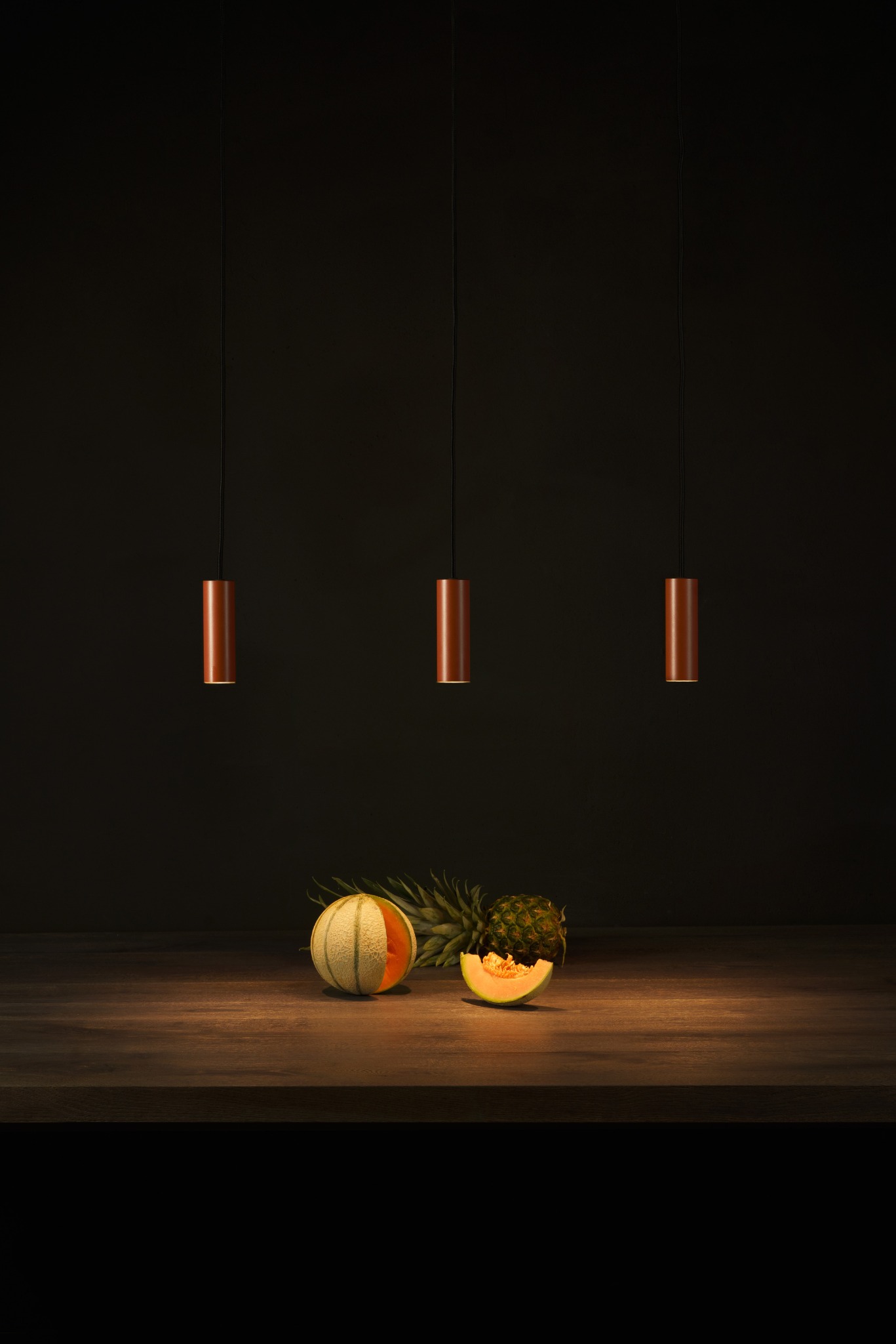 Trzy cylindryczne lampy wiszące w kolorze miedzi nad drewnianym blatem z melonem i ananasem. Minimalistyczny design oświetlenia w ciemnym otoczeniu.