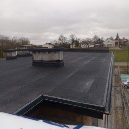 Grzejemy papę Rafał Wrona - Czarny dach płaski z kominami na tle zabudowy jednorodzinnej i pochmurnego nieba. Widoczne rusztowanie i opakowanie materiałów budowlanych.