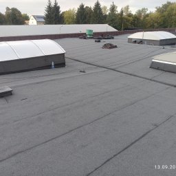 Grzejemy papę Rafał Wrona - Szary dach płaski z świetlikami dachowymi i pracownikiem w oddali. Widoczne detale łączeń papy i elementy wentylacyjne. Data wykonania zdjęcia w prawym dolnym rogu.