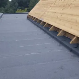 Szczegółowe ujęcie płaskiego dachu pokrytego papą, z widocznymi obróbkami blacharskimi i elementami konstrukcji drewnianej. Dach w trakcie prac wykończeniowych.