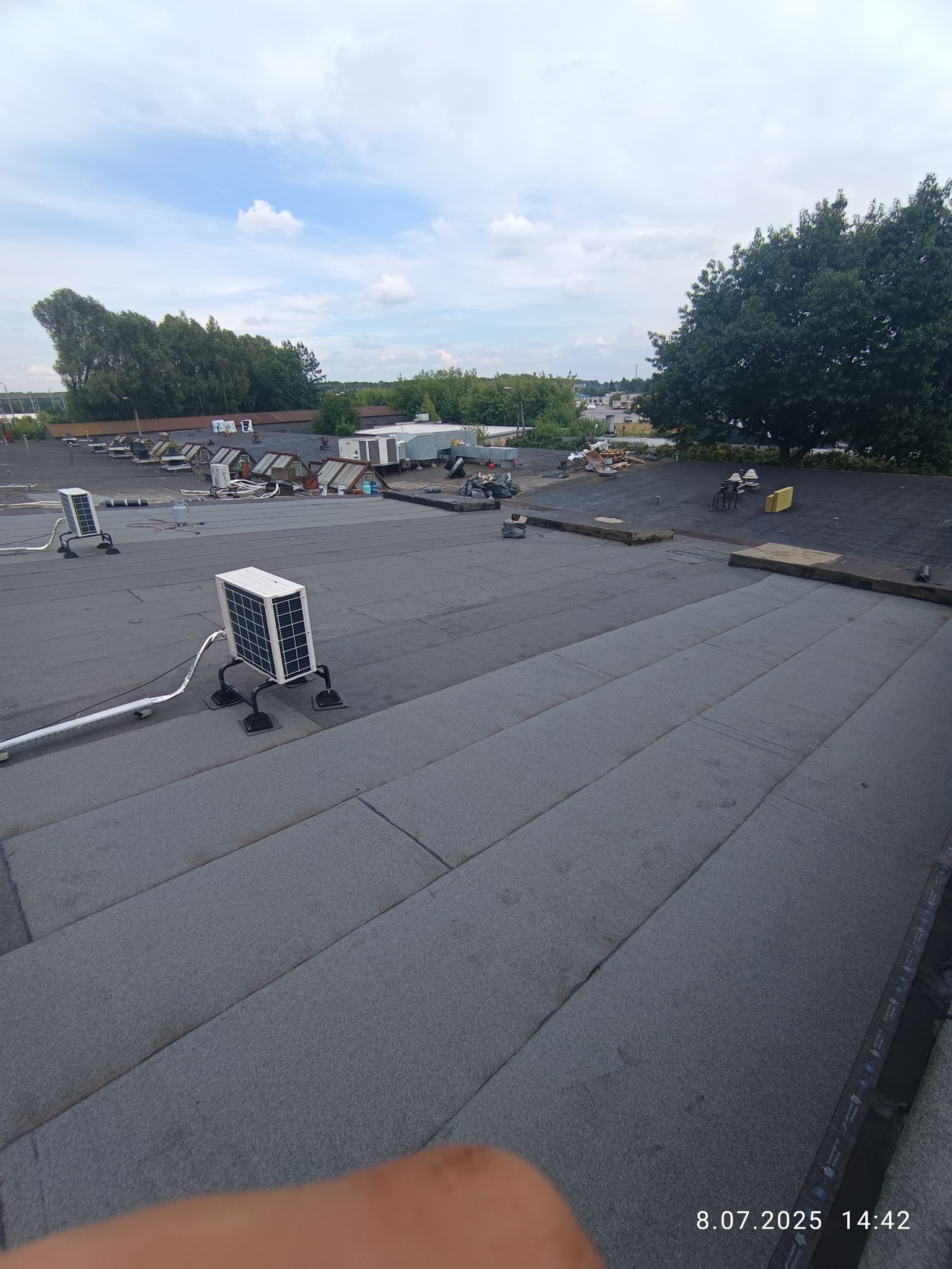 Szary dach pokryty papą z zamontowanymi klimatyzatorami. W tle widoczne budynki i drzewa. Ujęcie z góry, z widocznym fragmentem dłoni w dolnym rogu.