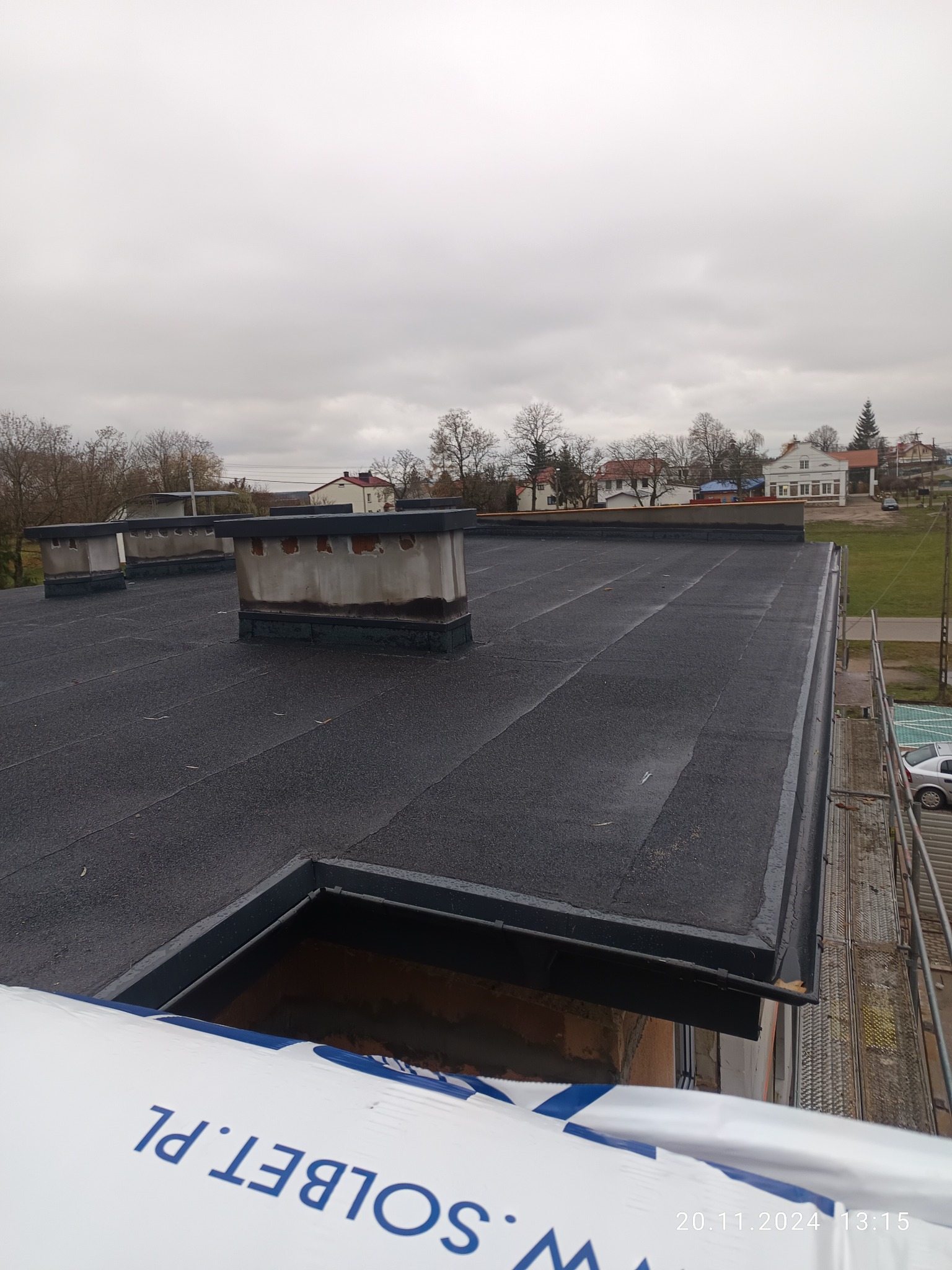 Czarny dach płaski z kominami na tle zabudowy jednorodzinnej i pochmurnego nieba. Widoczne rusztowanie i opakowanie materiałów budowlanych.