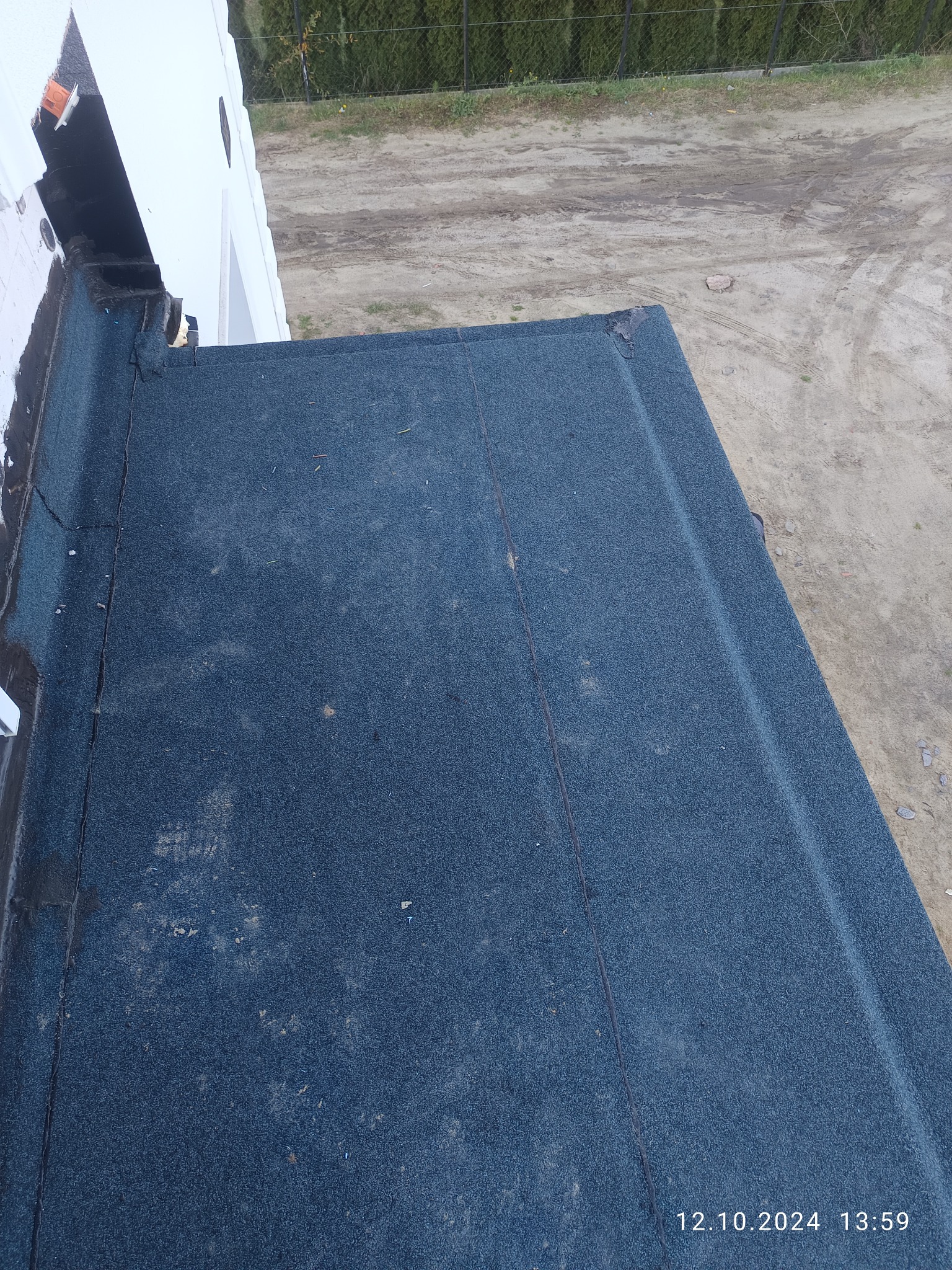 Fragment dachu pokrytego papą termozgrzewalną, widok z góry. Widoczne łączenia papy i fragment ściany budynku. W tle ogrodzenie i ziemia.