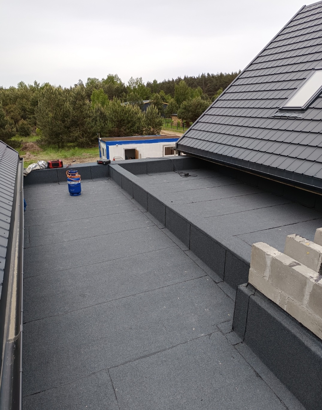 Wykończony płaski dach pokryty papą termozgrzewalną z detalami obróbek blacharskich i fragmentem dachu skośnego z szarą dachówką ceramiczną.