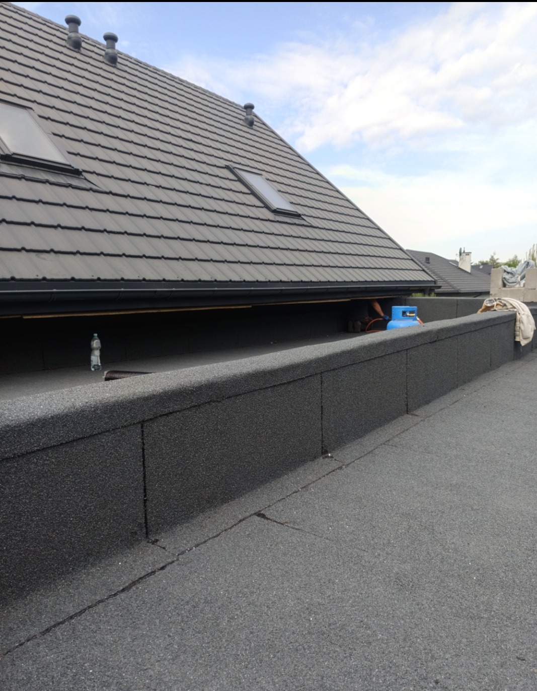 Czarny, płaski dach z widoczną konstrukcją i oknami dachowymi. W tle błękitne niebo z chmurami. Widoczny fragment prac dekarskich i butla gazowa.