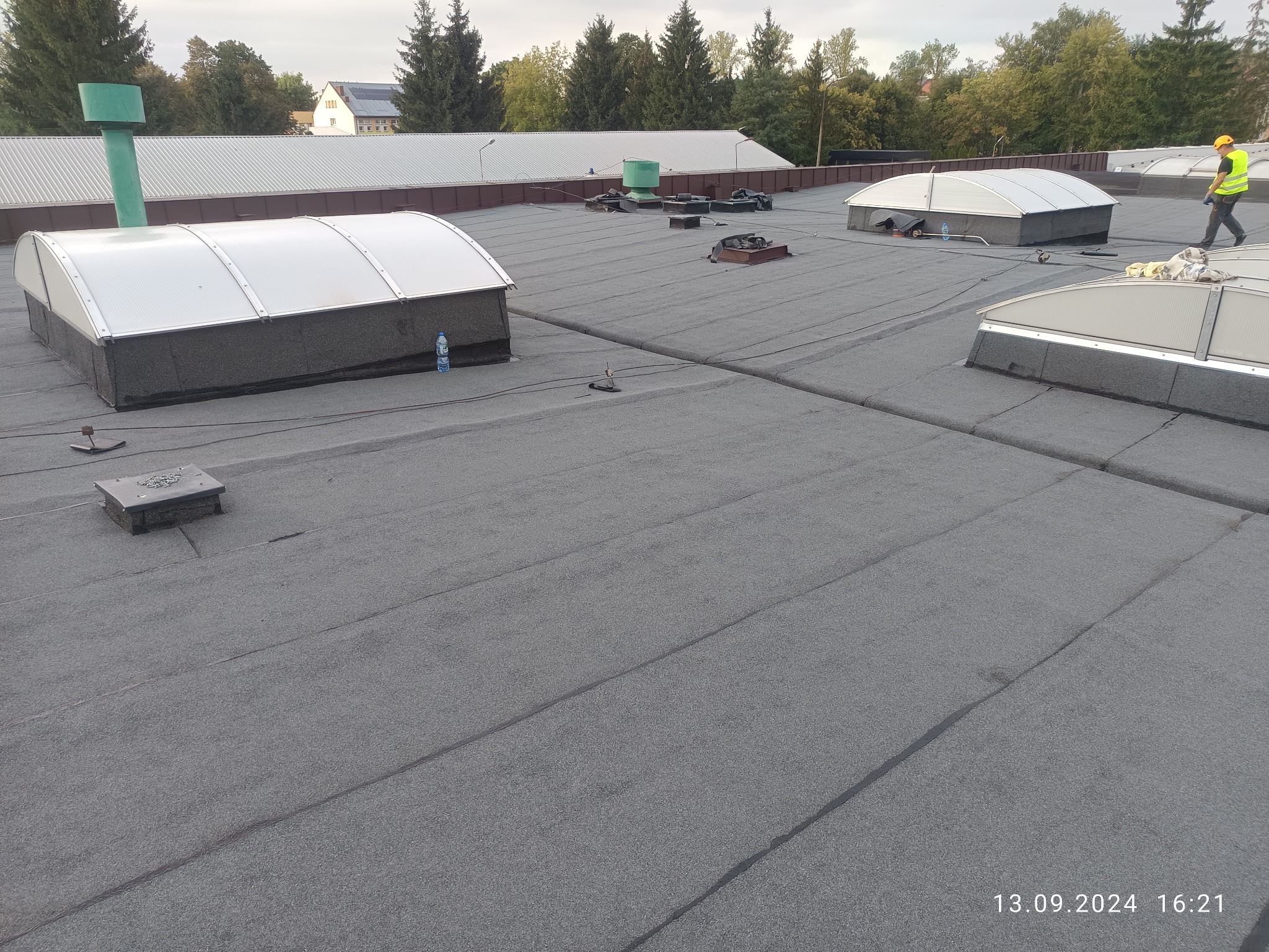 Szary dach płaski z świetlikami dachowymi i pracownikiem w oddali. Widoczne detale łączeń papy i elementy wentylacyjne. Data wykonania zdjęcia w prawym dolnym rogu.
