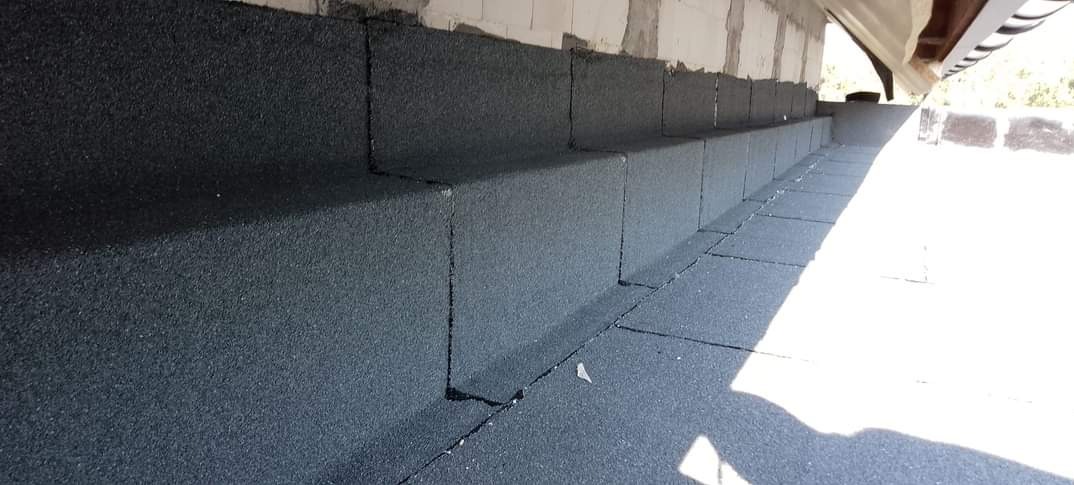 Szczegółowe ujęcie dachu pokrytego czarną, bitumiczną papą, ukazujące izolację przy ścianie budynku oraz fragment rynny w tle. Widoczne połączenia papy.