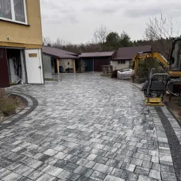 Nowa, szara kostka brukowa ułożona na podjeździe posesji z otwartym garażem i wjazdem, w tle widoczny minikoparka i zagęszczarka.
