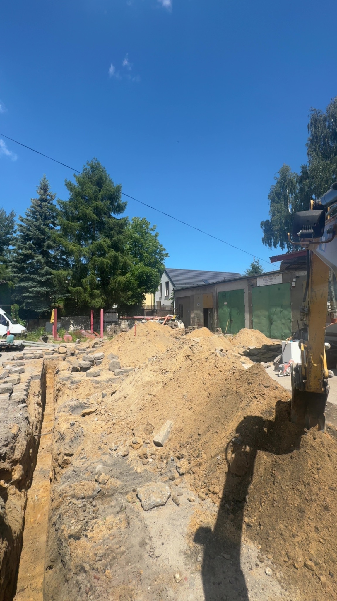 Wykopany rów z fragmentami kostki brukowej i stertą ziemi, obok widoczny fragment koparki. W tle budynek z zielonymi bramami garażowymi i błękitne niebo.