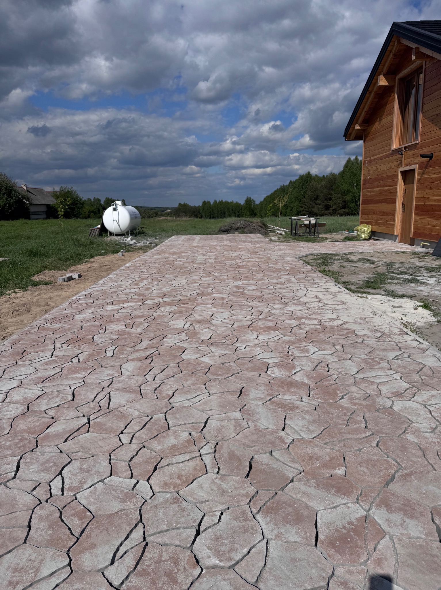 Ułożona ścieżka z nieregularnej kostki brukowej w odcieniach czerwieni, prowadząca do drewnianego domu w Skarżysku-Kamiennej. Tło: pochmurne niebo.