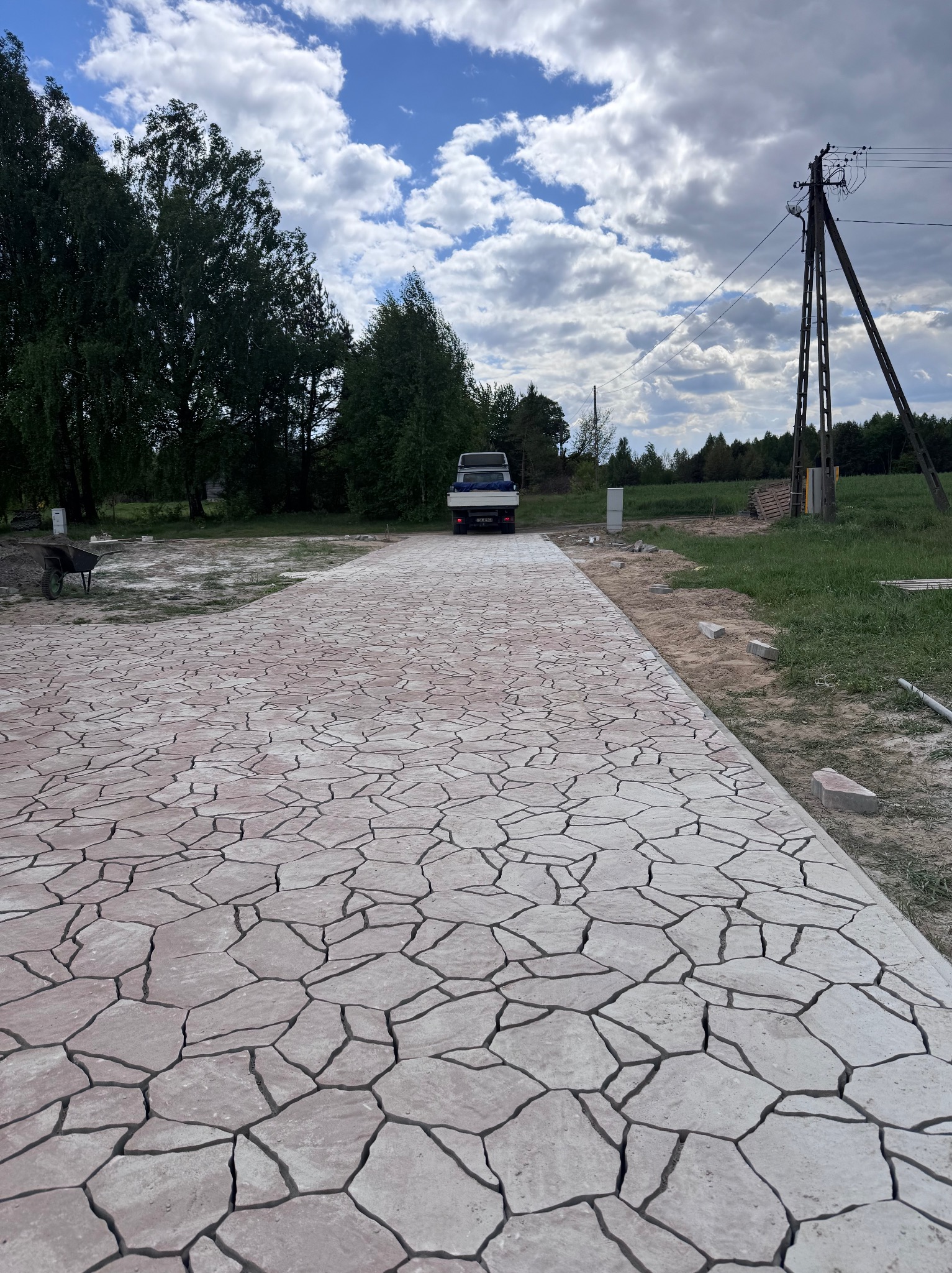 Ułożona ścieżka z nieregularnej kostki brukowej w odcieniach beżu i szarości, prowadząca w kierunku zaparkowanego samochodu dostawczego na tle zieleni.