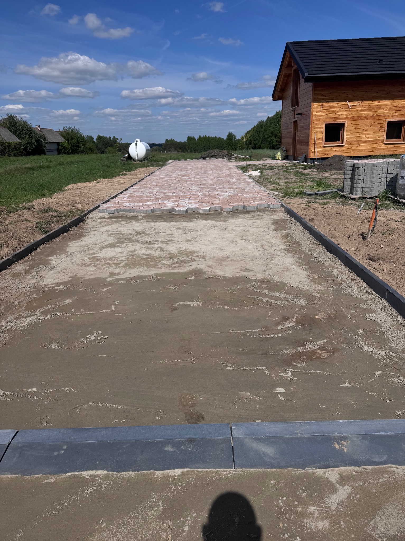 Częściowo ułożona, czerwono-szara kostka brukowa na ścieżce z obrzeżami. W tle dom z drewna i zbiornik na gaz. Widok z góry na plac budowy.