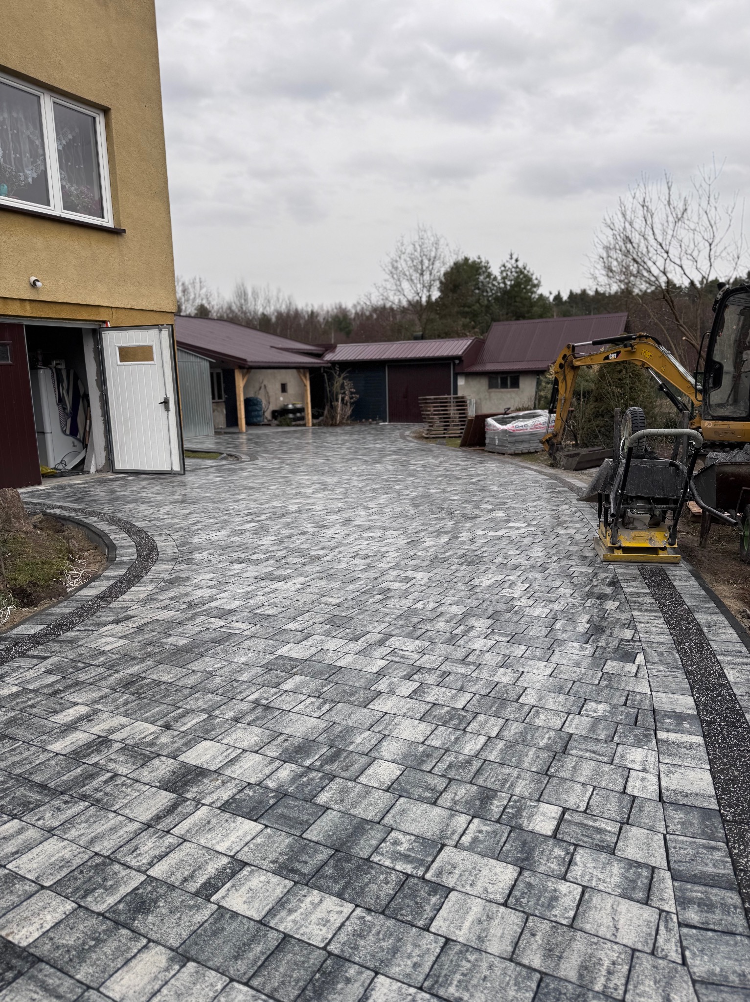 Nowa, szara kostka brukowa ułożona na podjeździe posesji z otwartym garażem i wjazdem, w tle widoczny minikoparka i zagęszczarka.