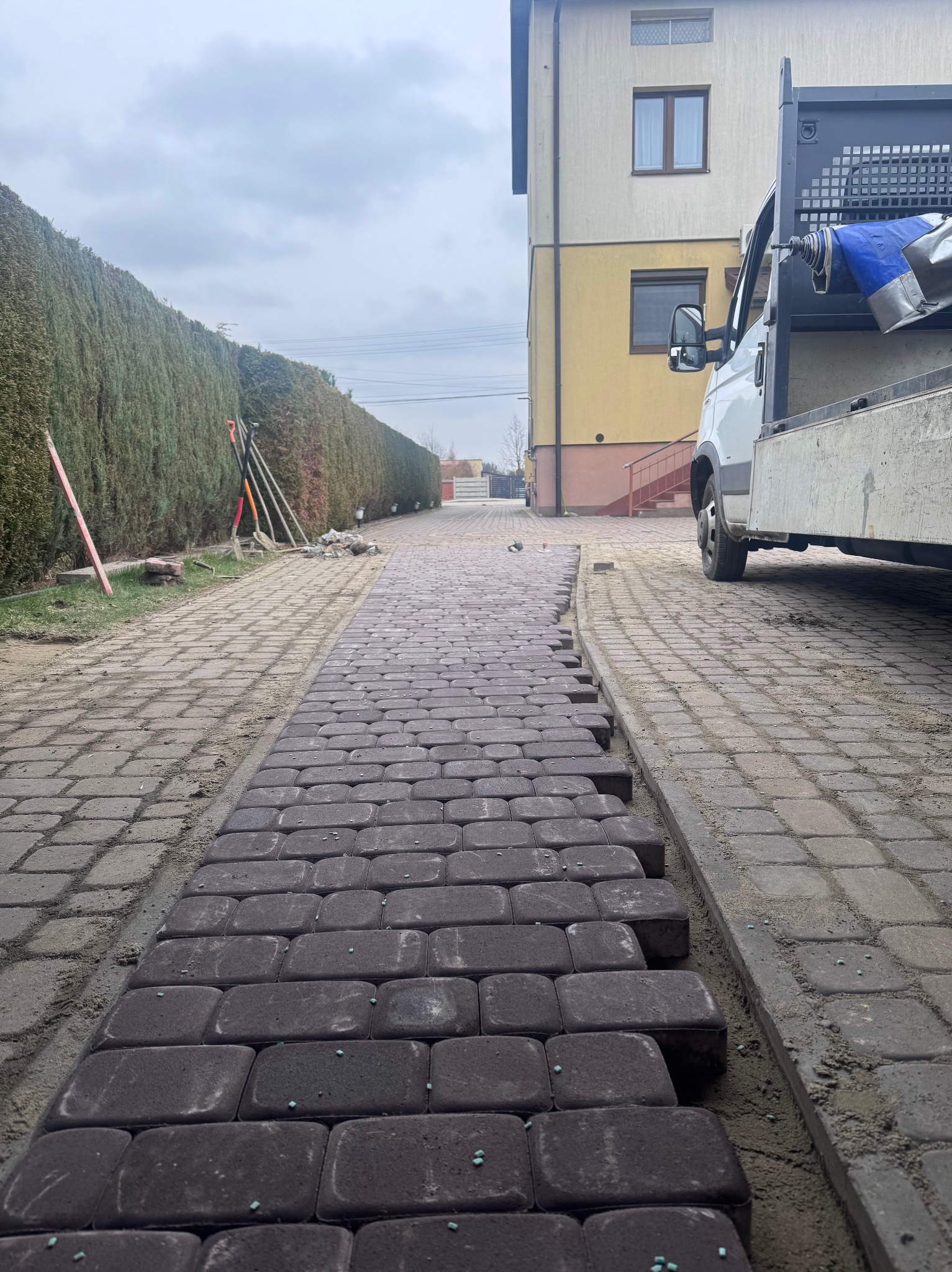 Częściowo ułożona, grafitowa kostka brukowa na ścieżce, obok żywopłot i zaparkowany samochód dostawczy. Widoczne narzędzia brukarskie.
