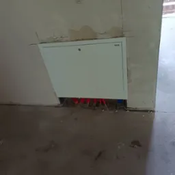 Biała skrzynka rewizyjna TECE z widocznymi czerwonymi rurami instalacji grzewczej w surowym wnętrzu. Fragment ściany zarysowany ołówkiem, surowa podłoga.