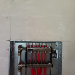 Rozdzielacz ogrzewania podłogowego TECE w metalowej szafce, zamontowany na surowej ścianie z widocznymi oznaczeniami instalacyjnymi. Czerwone rury i zawory.