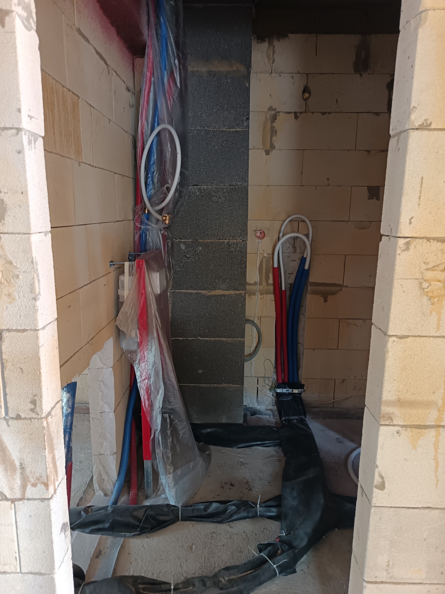 Instalacja rur wodnych (czerwone i niebieskie) w surowym wnętrzu, widoczne bloczki i folia ochronna. Etap prac instalacyjnych w nowym budynku.
