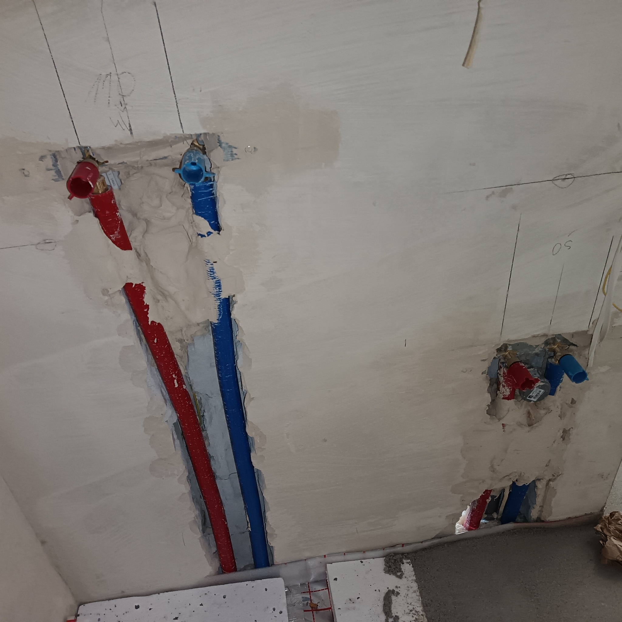 Ściana z instalacją wodną: czerwone i niebieskie rury w bruzdach, częściowo zamaskowane gipsem. Widoczne złącza i fragment ogrzewania podłogowego.