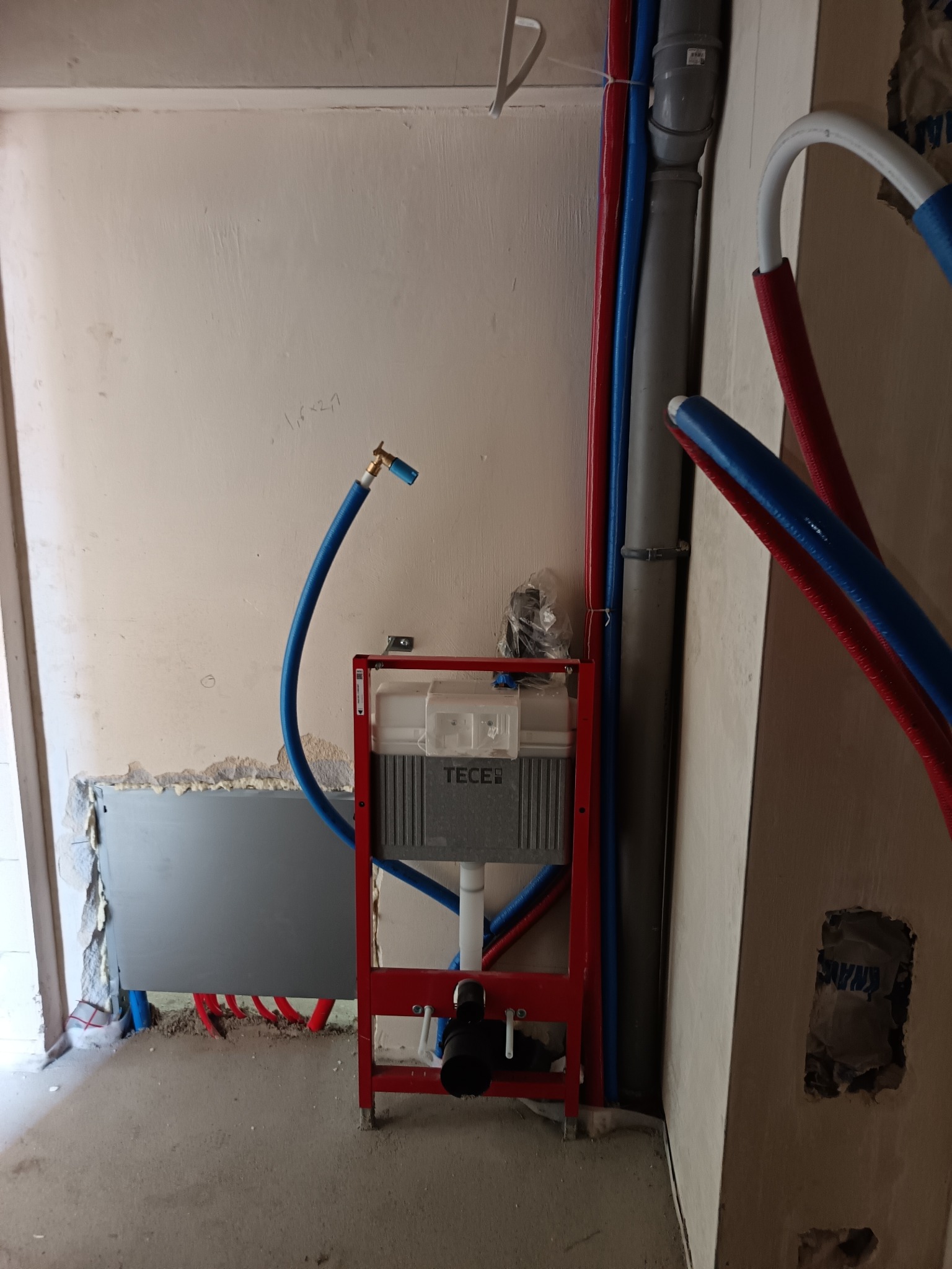 Montaż stelaża podtynkowego TECE z niebieską rurą doprowadzającą wodę i fragmentem instalacji grzewczej z czerwonymi i niebieskimi rurami w surowym wnętrzu.
