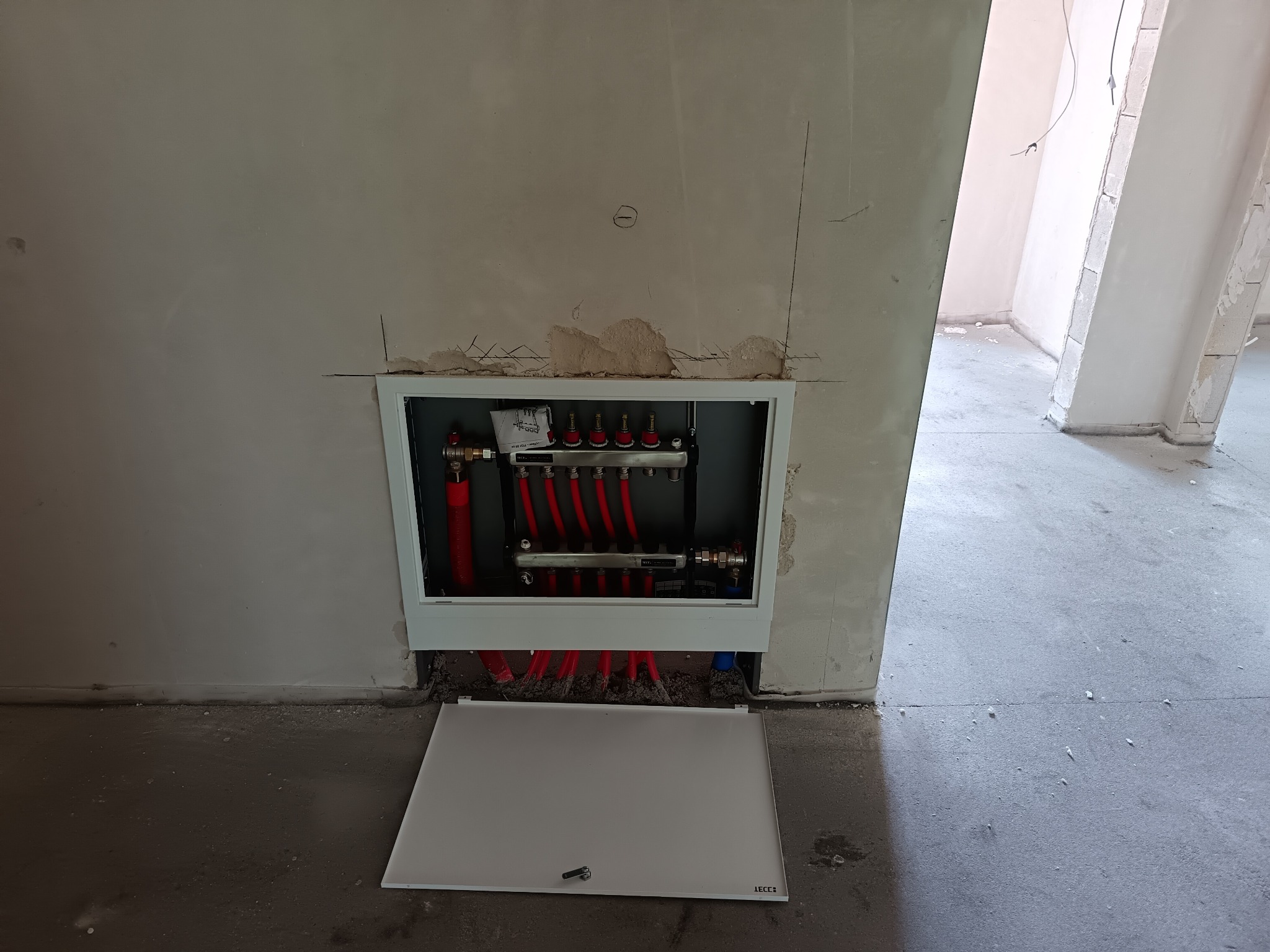 Rozdzielacz ogrzewania podłogowego w zabudowie, z widocznymi czerwonymi rurami i otwartymi drzwiczkami rewizyjnymi. Surowe ściany i podłoga w tle.