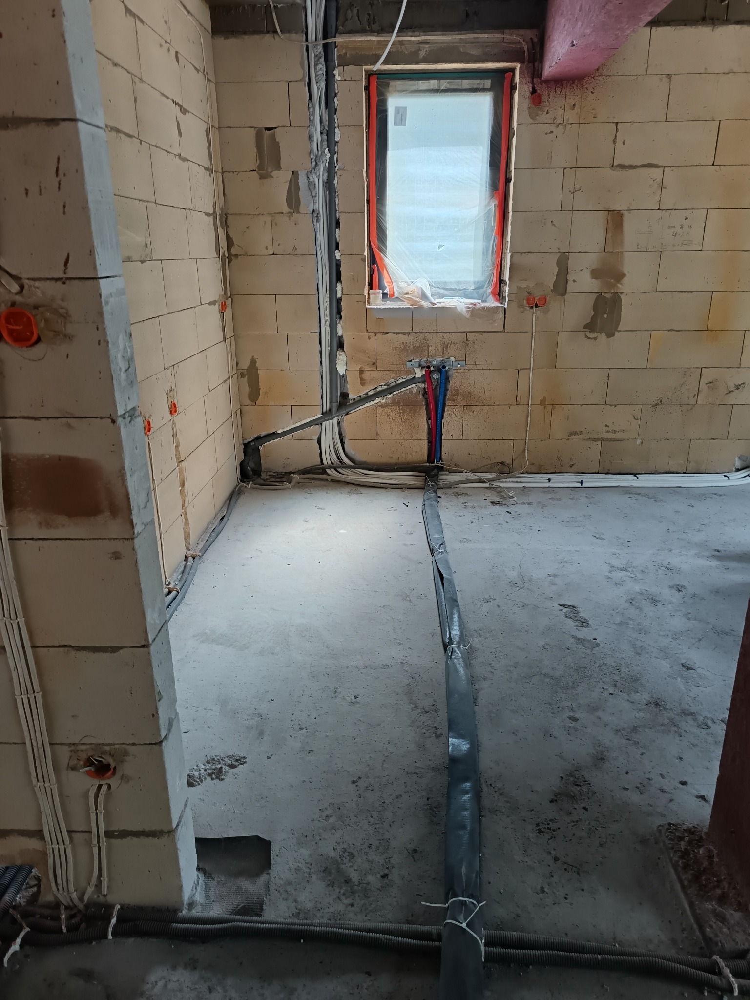 Surowy stan: instalacja elektryczna i hydrauliczna w nowym budynku. Widoczne przewody, rury (czerwona i niebieska), puszki w ścianach z betonu komórkowego. Okno z folią.