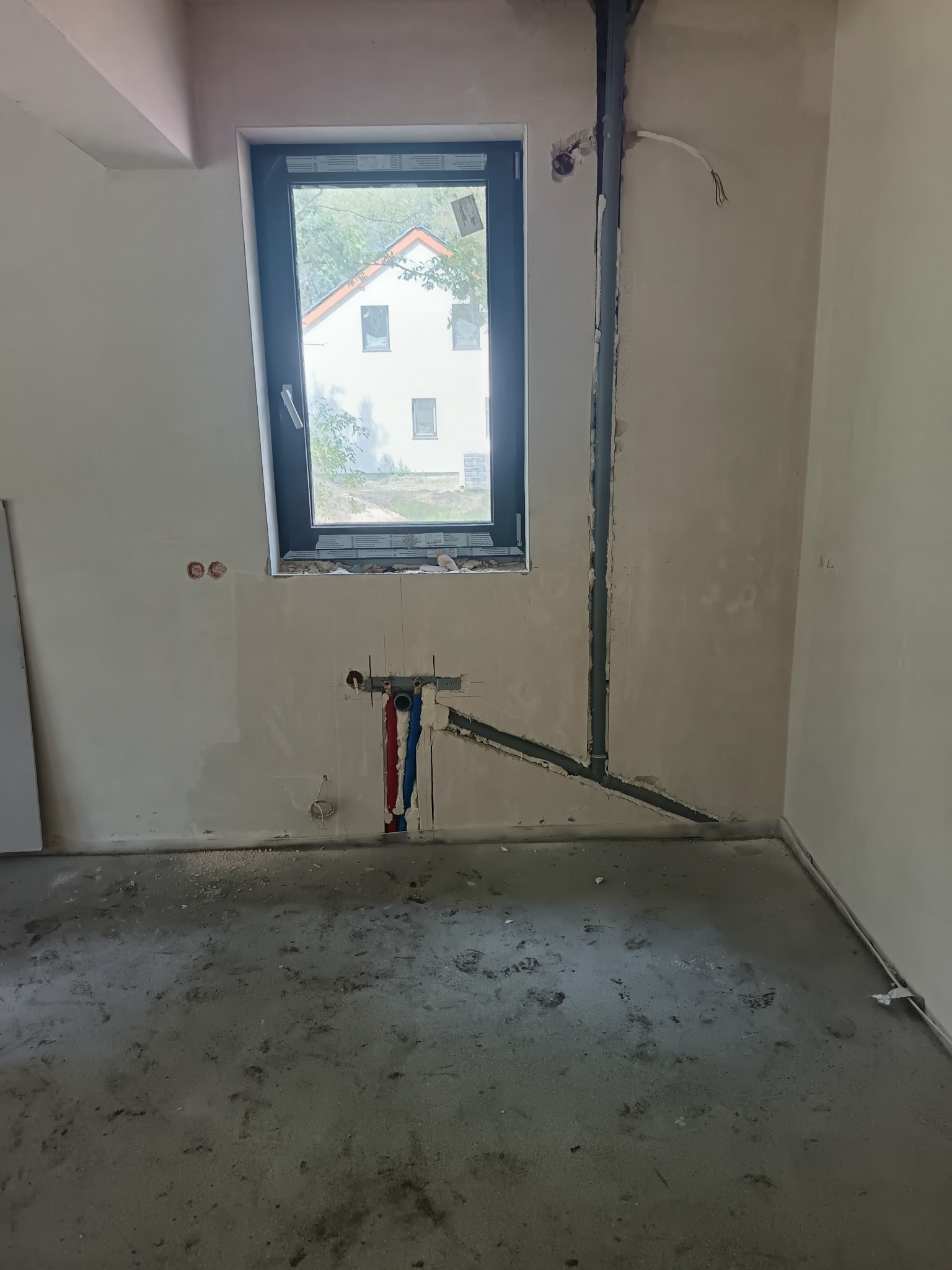 Wnętrze w trakcie prac instalacyjnych. Widoczne rury instalacji wodnej (czerwona i niebieska) oraz szara rura pionowa. W tle okno z widokiem na dom.