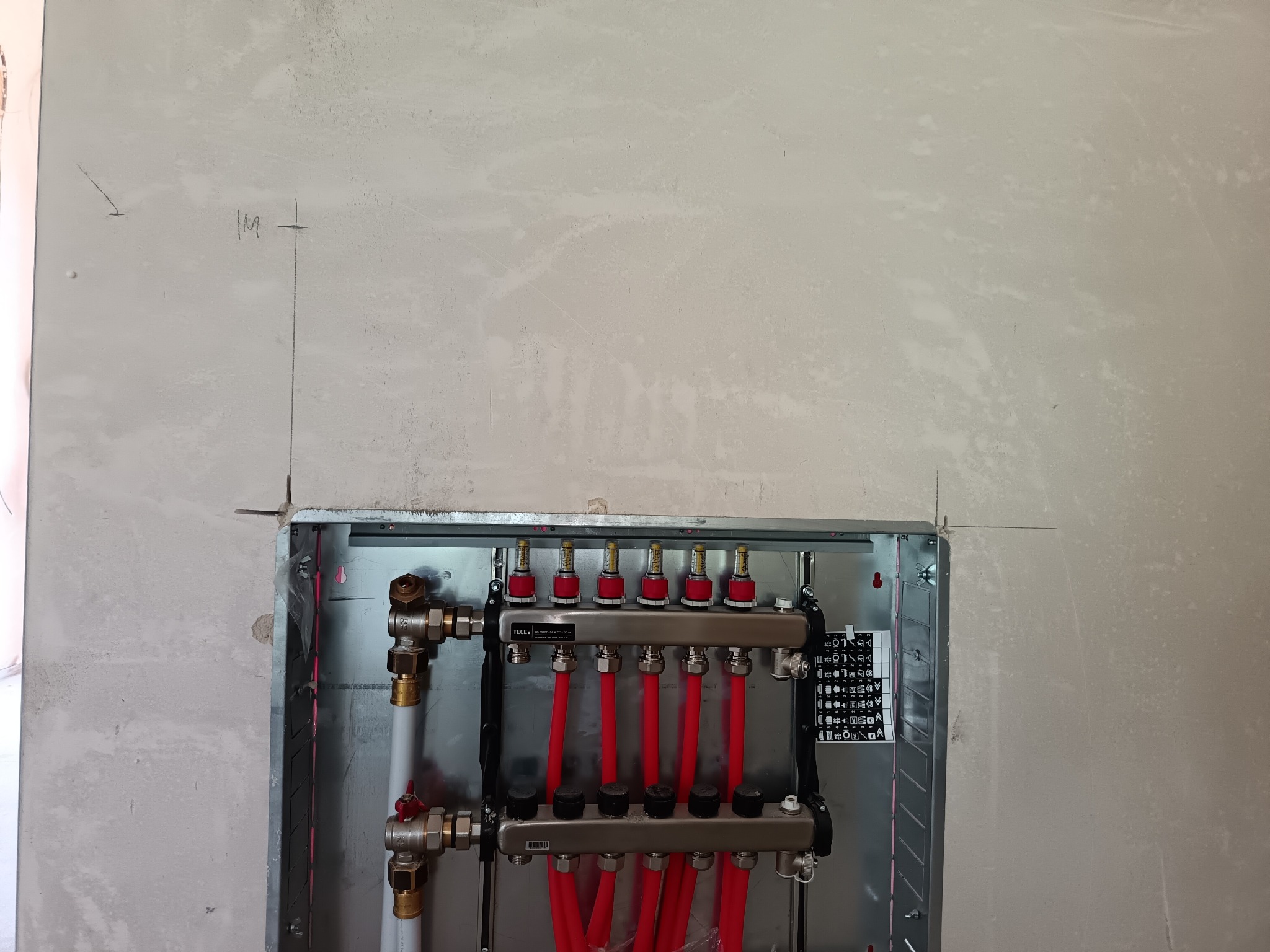Rozdzielacz ogrzewania podłogowego TECE w metalowej szafce, zamontowany na surowej ścianie z widocznymi oznaczeniami instalacyjnymi. Czerwone rury i zawory.