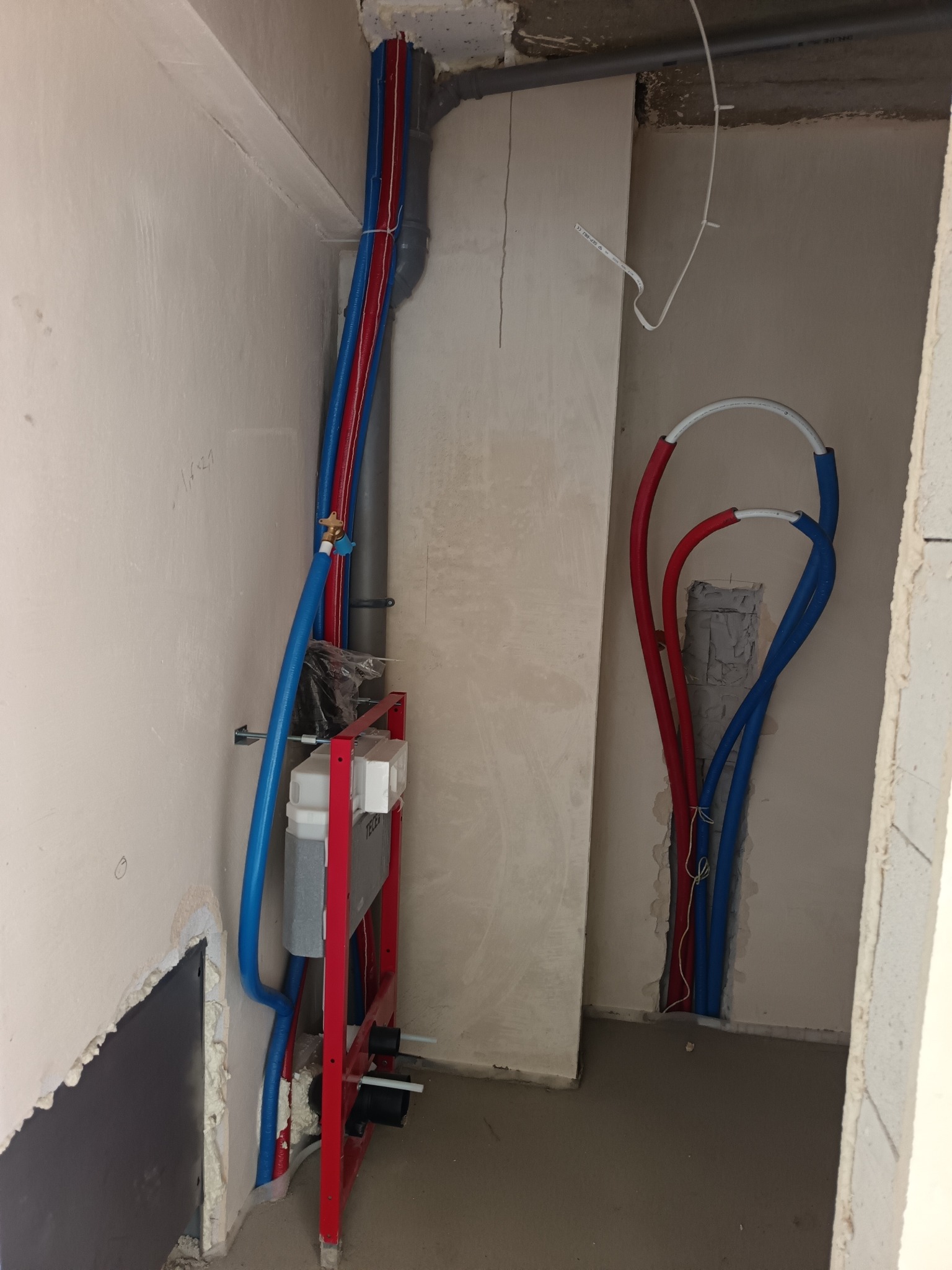 Montaż instalacji wodnej: czerwone i niebieskie rury prowadzone wzdłuż ściany, instalacja podtynkowa WC na metalowym stelażu, surowy stan.