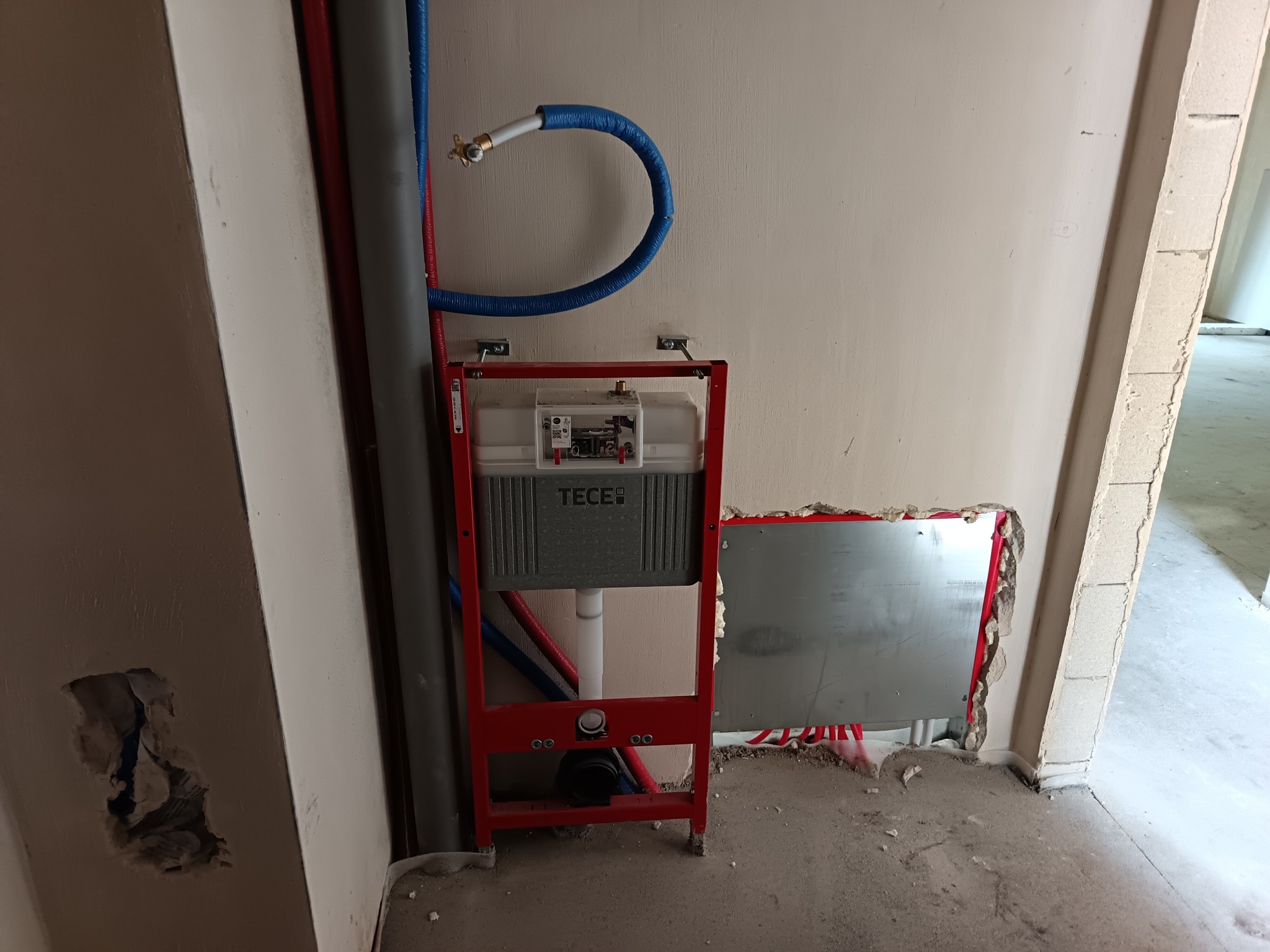 Stelaż podtynkowy TECE z niebieską rurą doprowadzającą wodę, zamontowany przy ścianie w surowym stanie, widoczne rury instalacyjne w kolorze czerwonym i szarym.