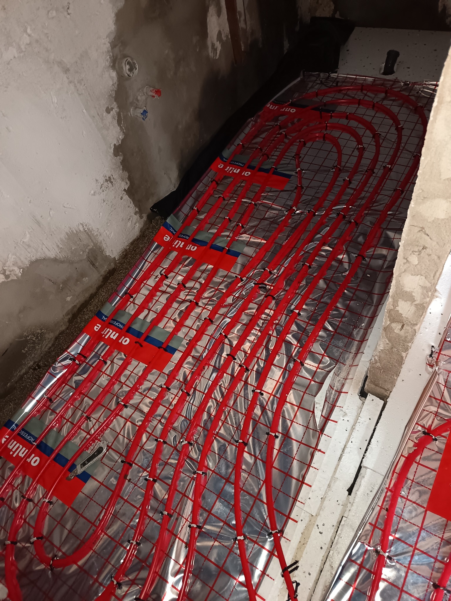 Czerwone rury ogrzewania podłogowego ułożone na srebrnej folii izolacyjnej, przymocowane czarnymi klipsami, widoczne surowe ściany w tle.