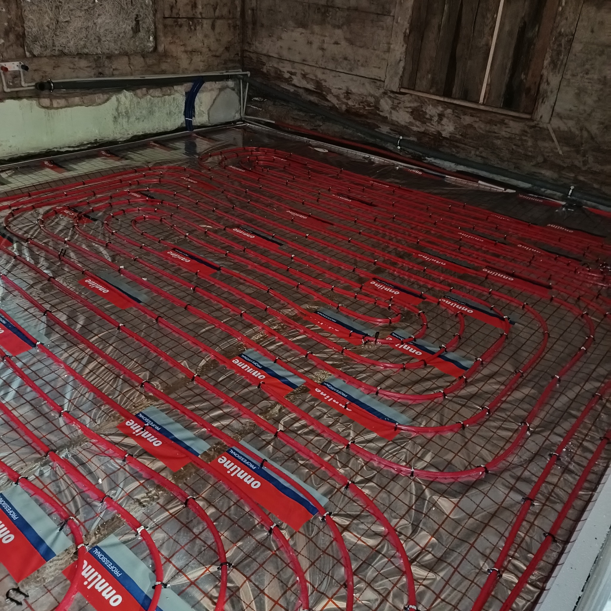 Czerwone rury ogrzewania podłogowego ułożone na folii z siatką w pomieszczeniu o surowych ścianach i drewnianym oknie. Widok z góry na instalację przed zalaniem posadzki.