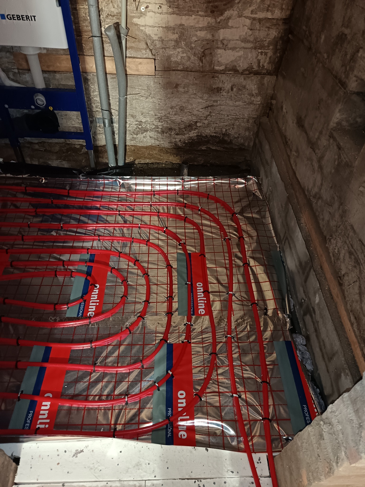 Czerwone rury ogrzewania podłogowego ułożone na folii izolacyjnej w surowym wnętrzu z betonowymi ścianami i drewnianym stropem. Widoczne elementy instalacji sanitarnej.