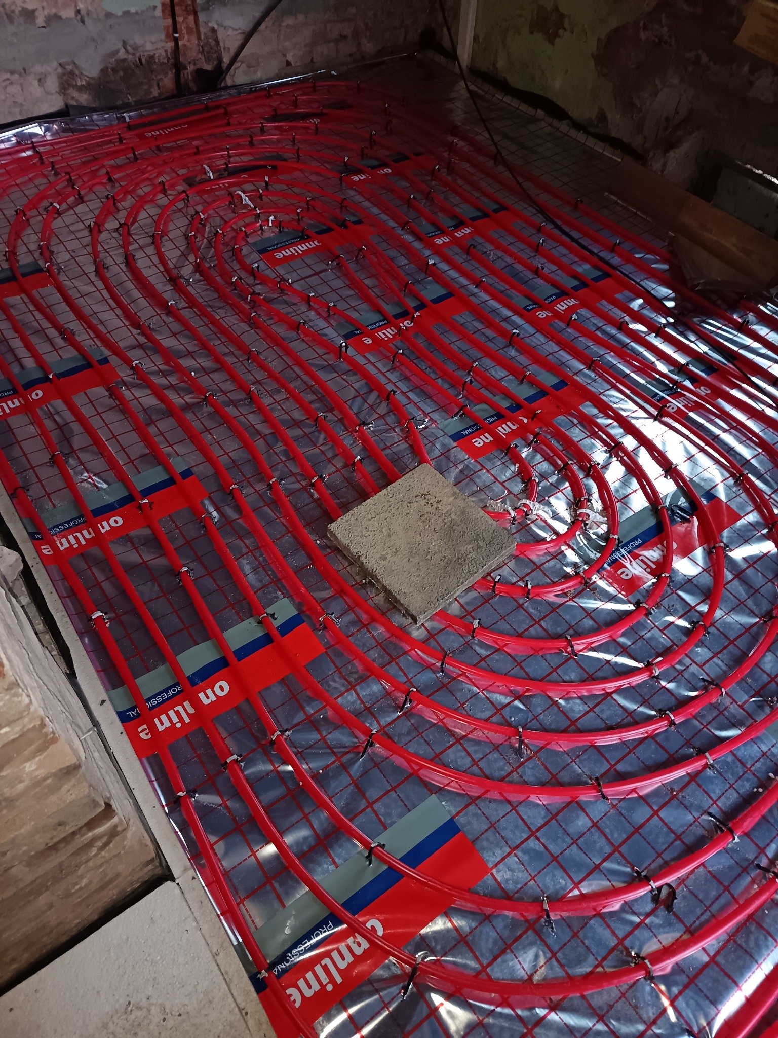 Czerwone rury ogrzewania podłogowego ułożone spiralnie na srebrnej folii z siatką, zabezpieczone czarnymi klipsami. Na środku leży szary bloczek.