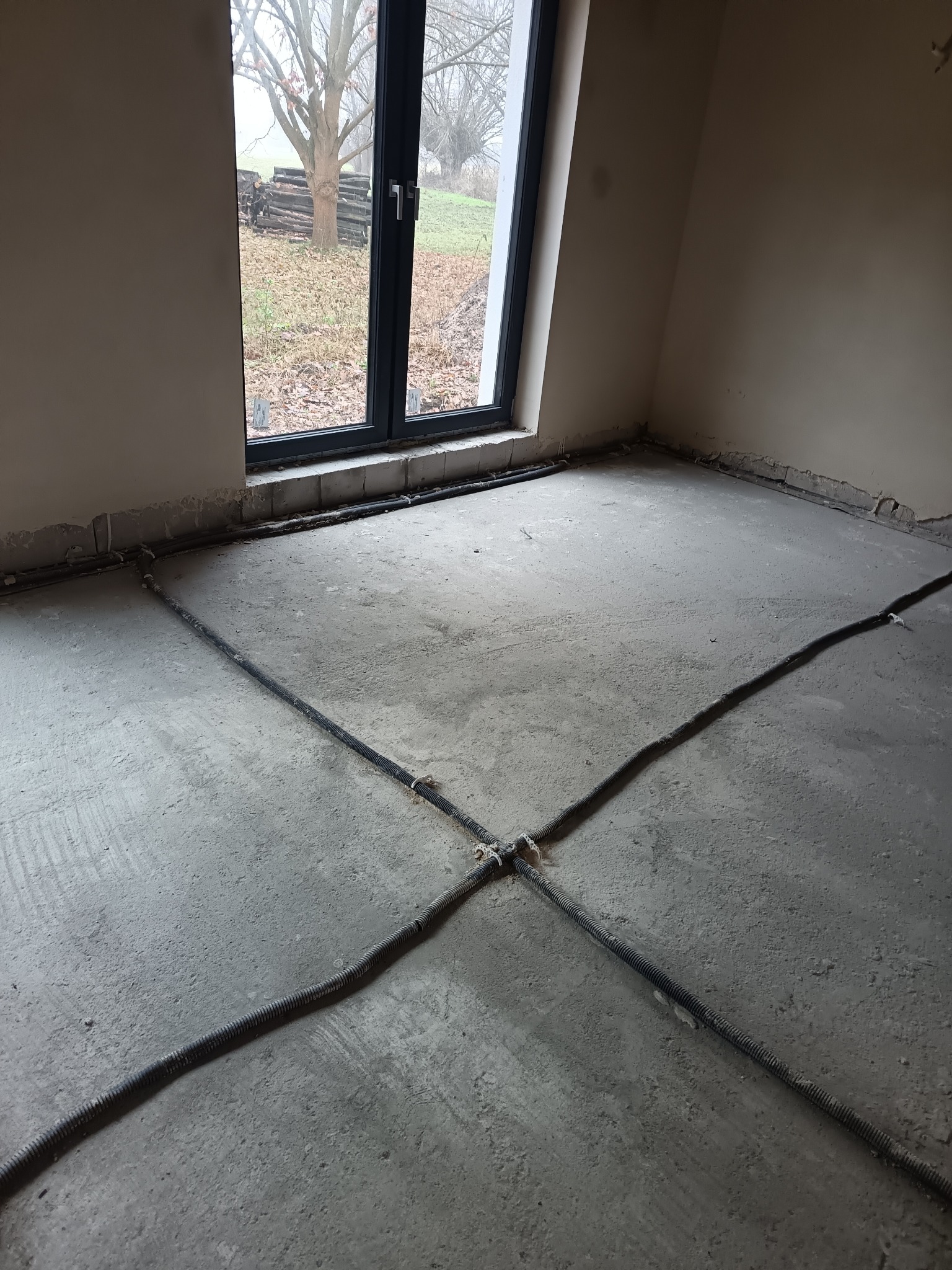 Szare rury w kształcie krzyża na betonowej podłodze w surowym wnętrzu z oknem. Instalacja ogrzewania podłogowego przed wylaniem posadzki.