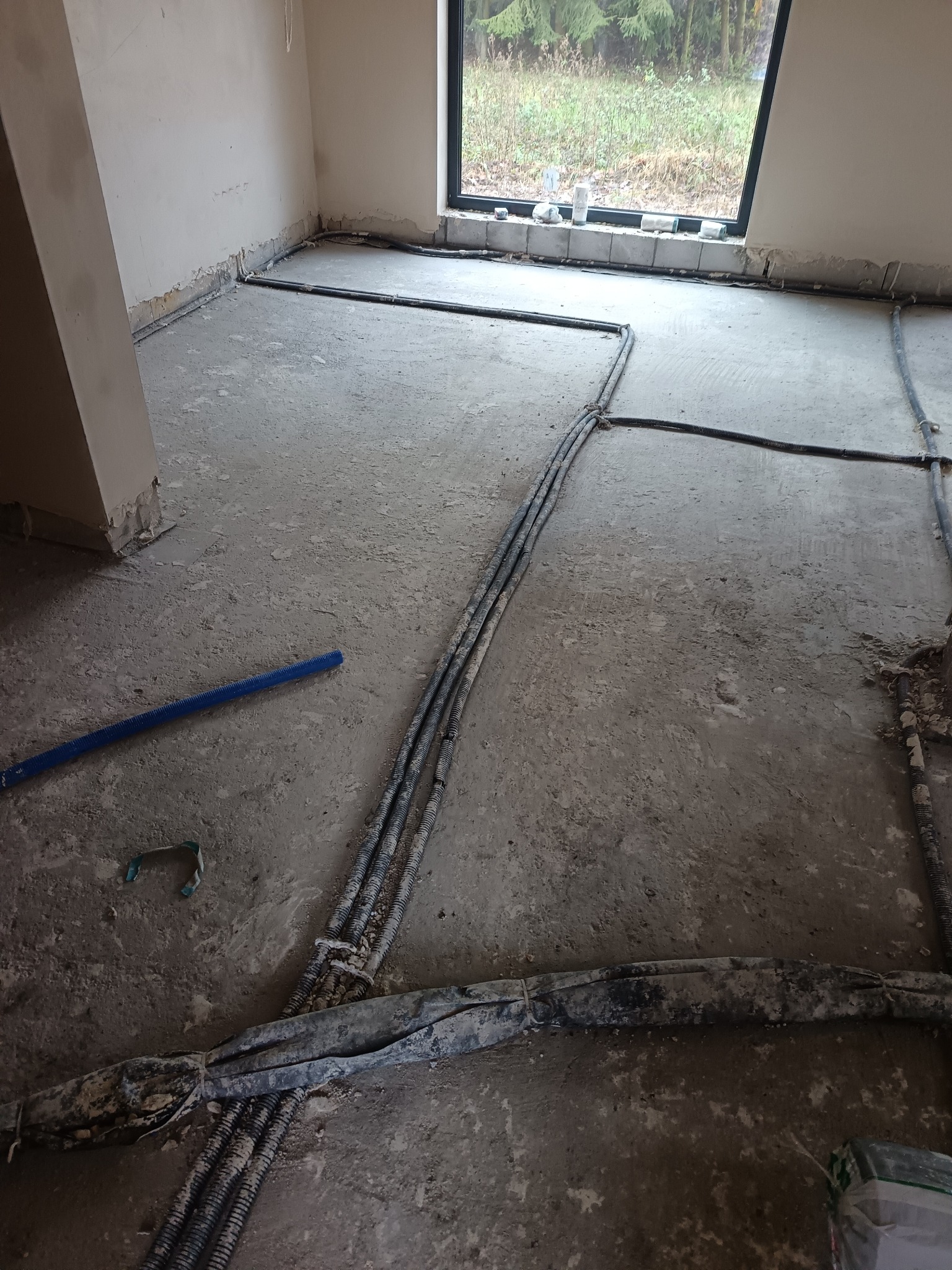 Szare rury instalacji grzewczej ułożone na surowym betonie podłogi w jasnym pomieszczeniu z oknem. Widoczne elementy mocujące i niebieska rura. Etap prac przed wylaniem posadzki.