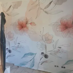 Ściana z motywem kwiatowym, częściowo zabezpieczona folią malarską, widoczne gniazdka elektryczne i rolka papieru. Dekoracyjne malowanie ścian w mieszkaniu.