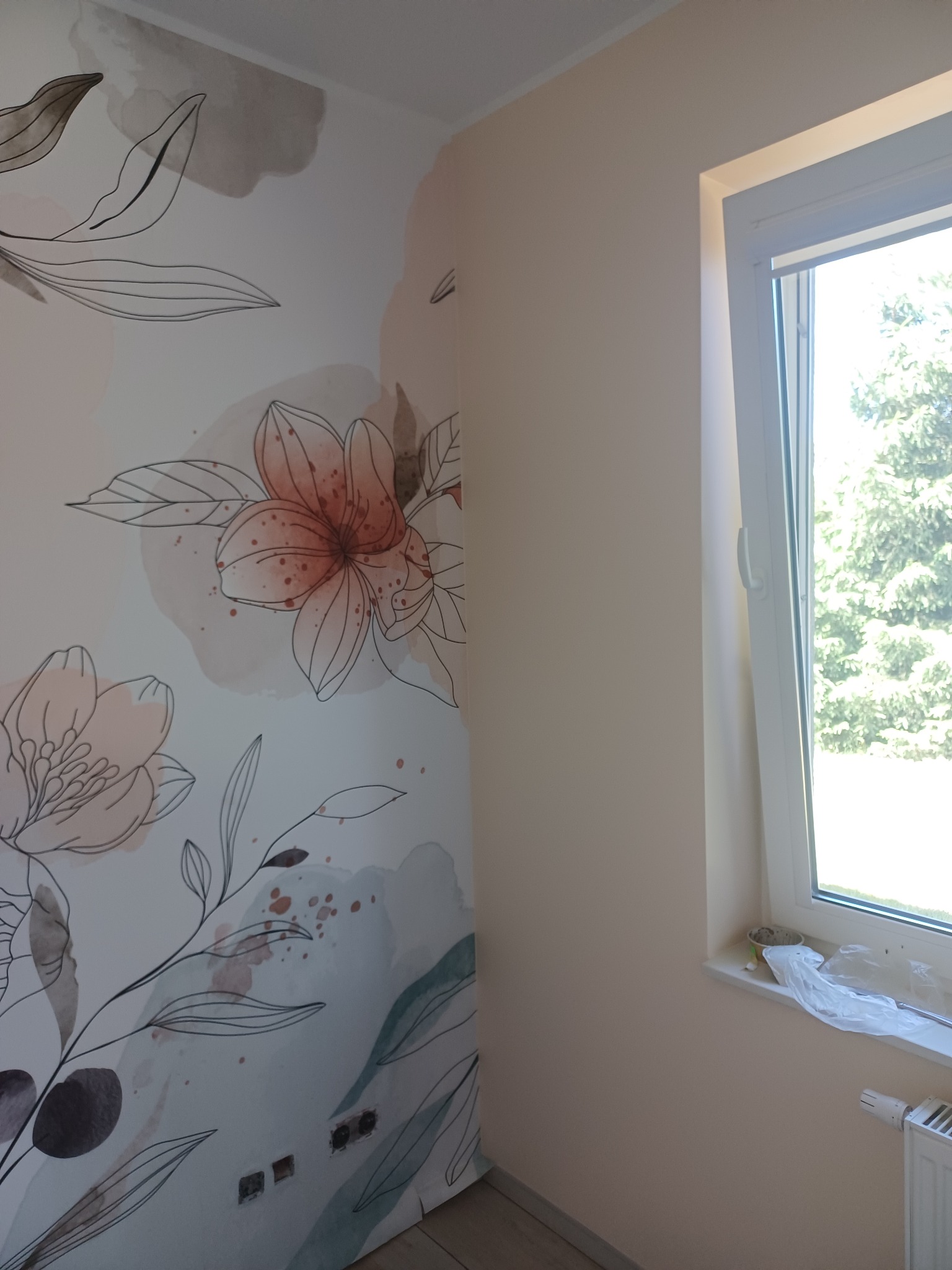 Ściana w pokoju po malowaniu z motywem kwiatowym, obok okno z widokiem na zieleń. Na parapecie resztki farby i folia malarska.
