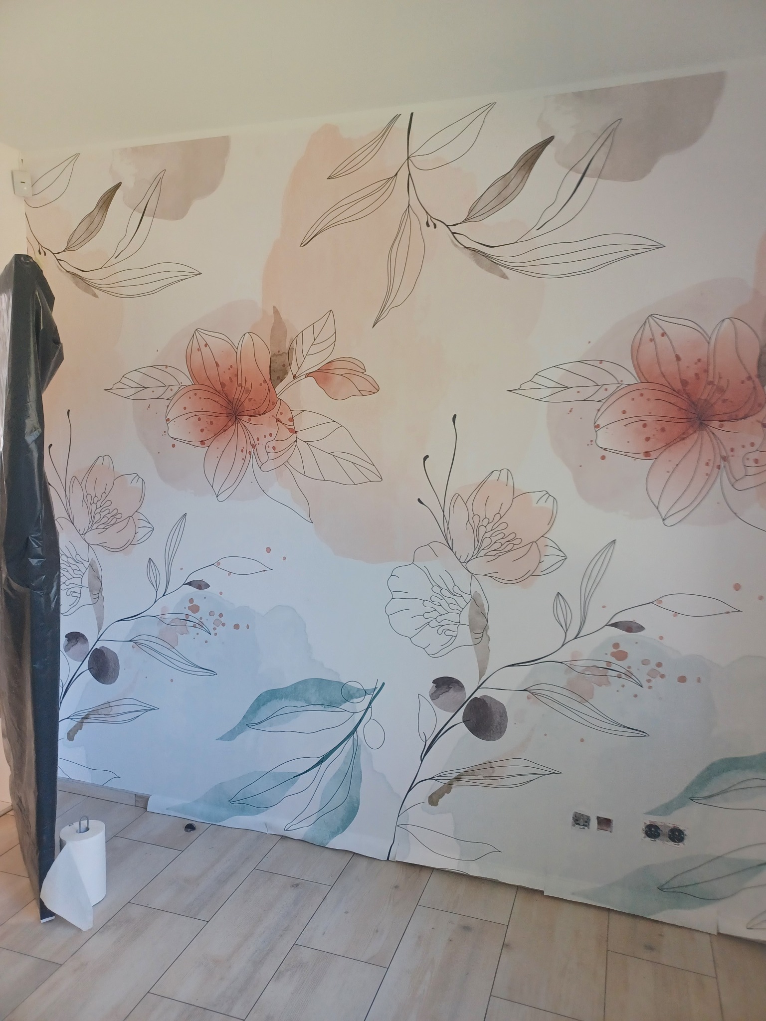 Ściana z motywem kwiatowym, częściowo zabezpieczona folią malarską, widoczne gniazdka elektryczne i rolka papieru. Dekoracyjne malowanie ścian w mieszkaniu.