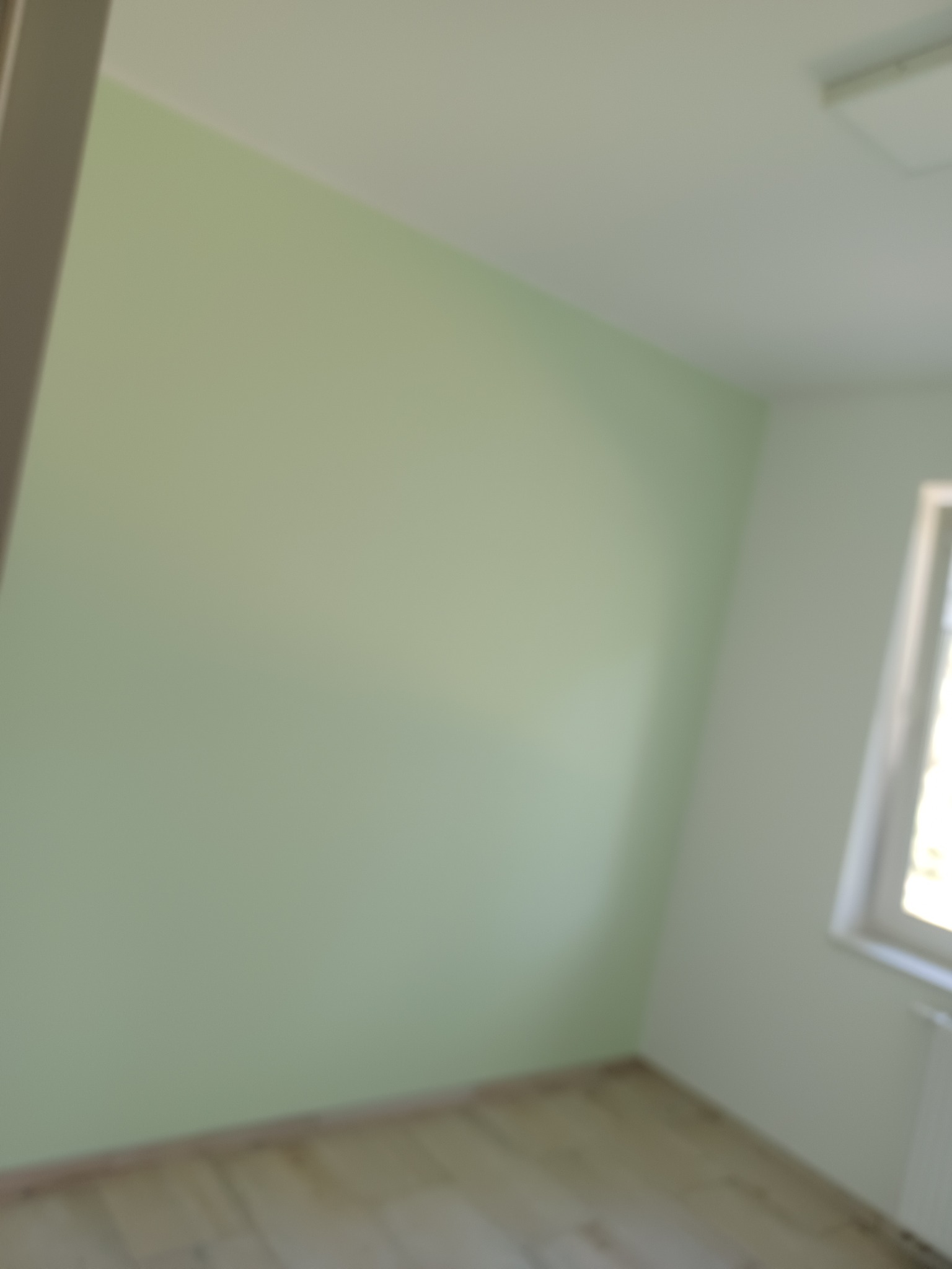 Świeżo pomalowany pokój z drewnianą podłogą i oknem w białej ramie. Ściana w odcieniach zieleni. Widoczne fragmenty grzejnika i sufitu.