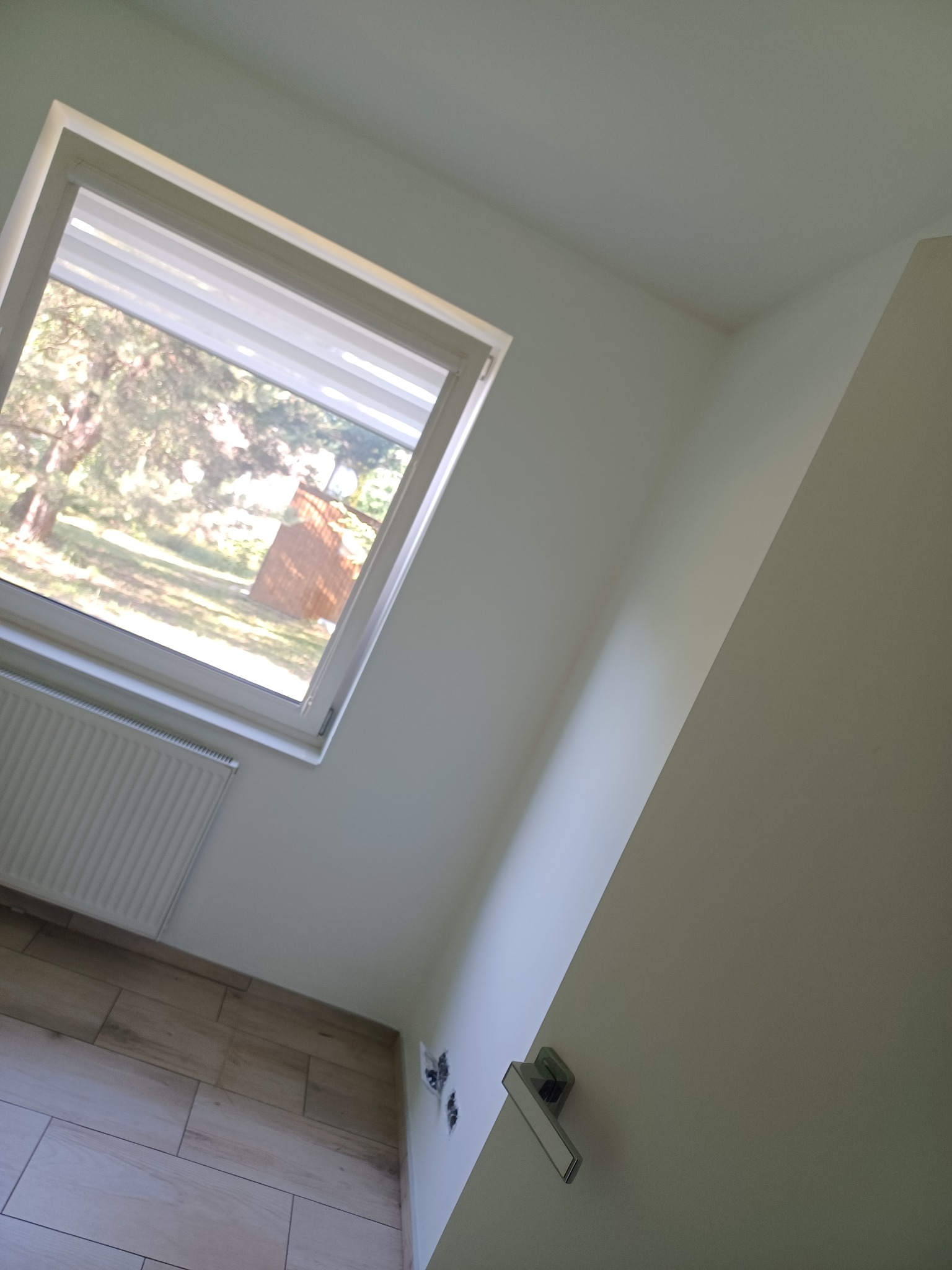 Jasne wnętrze po remoncie: świeżo pomalowane ściany w odcieniach bieli, okno z widokiem na zieleń, grzejnik i drewnopodobna podłoga. Minimalistyczny design.