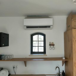 AIR Masters - Biały klimatyzator ścienny nad oknem w kuchni z drewnianym blatem i zlewem. Widoczna drabina i naczynia. Minimalistyczny design, jasne wnętrze.